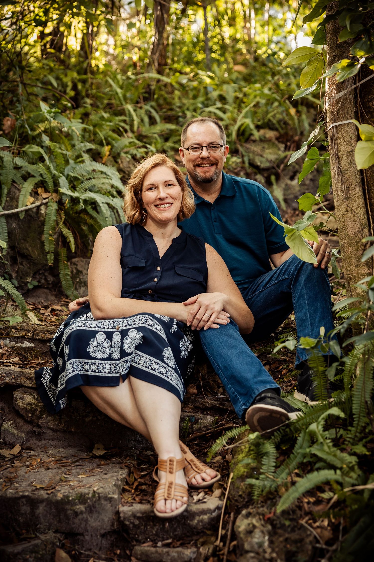 Smiling couple sitting on stone steps surrounded by ferns and greenery in a natural outdoor setting.