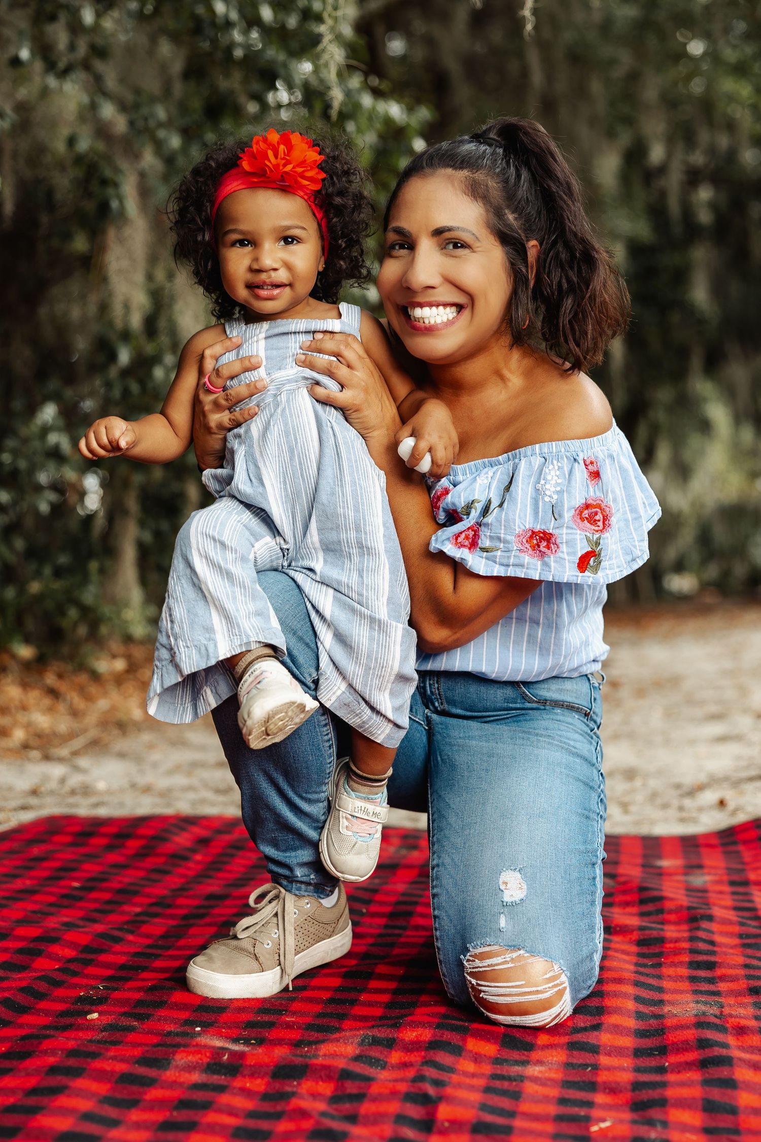 A smiling mother kneels on a red plaid blanket holding her toddler daughter outdoors.