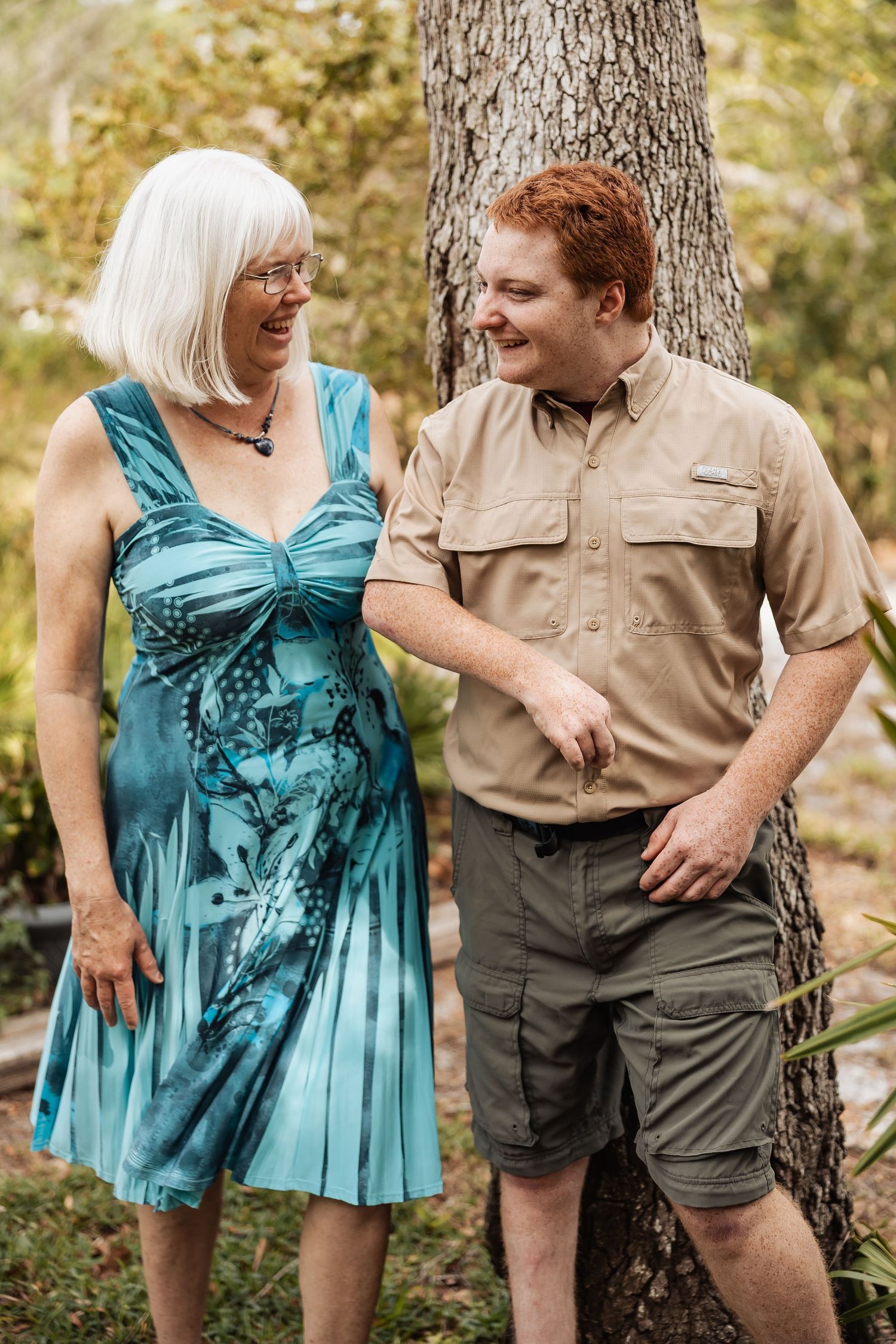 An older woman in a teal dress and a young redheaded man in khaki laugh together outdoors near a tree.