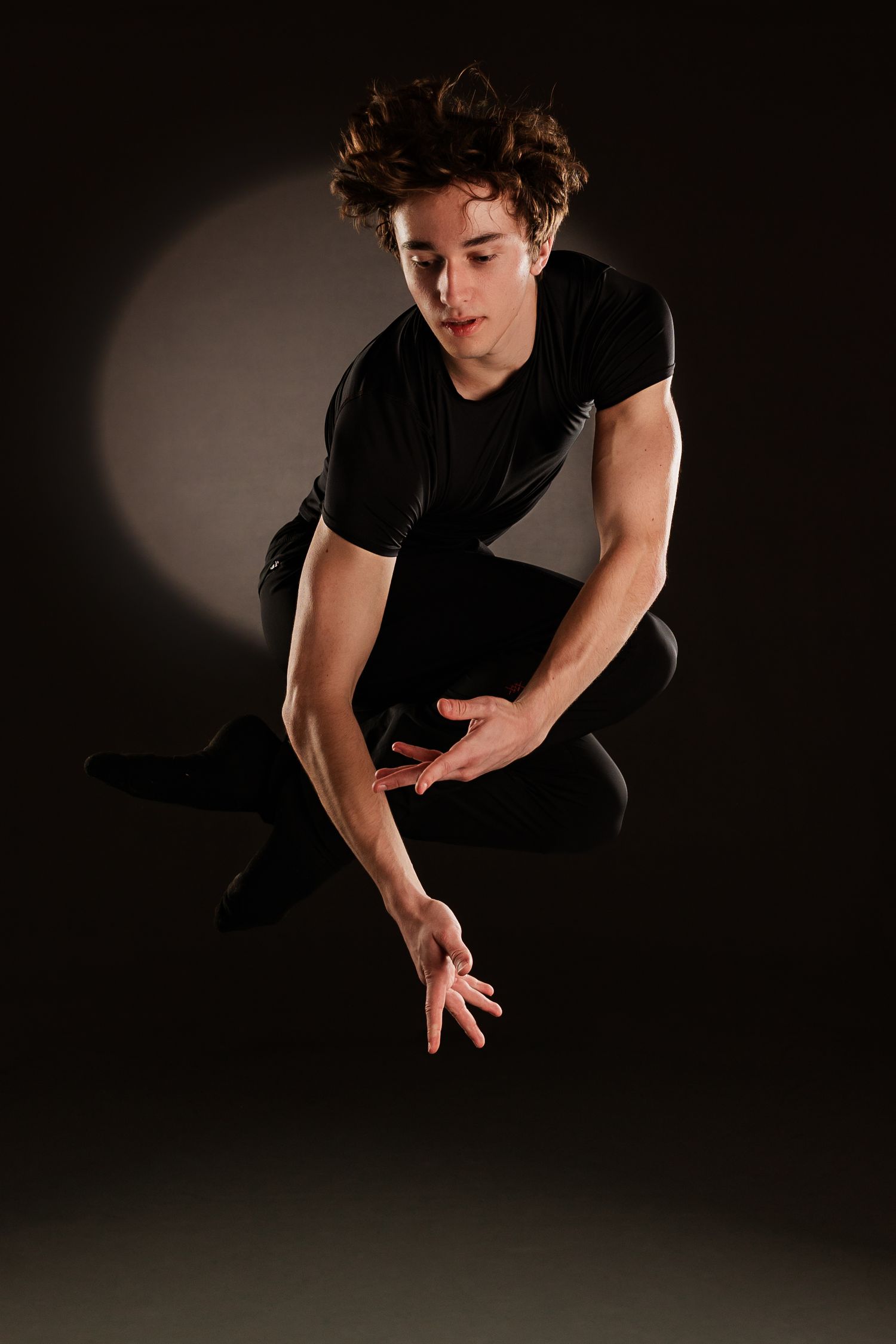 Young male dancer in black outfit mid-air, crouching with hands extended forward against dark background.