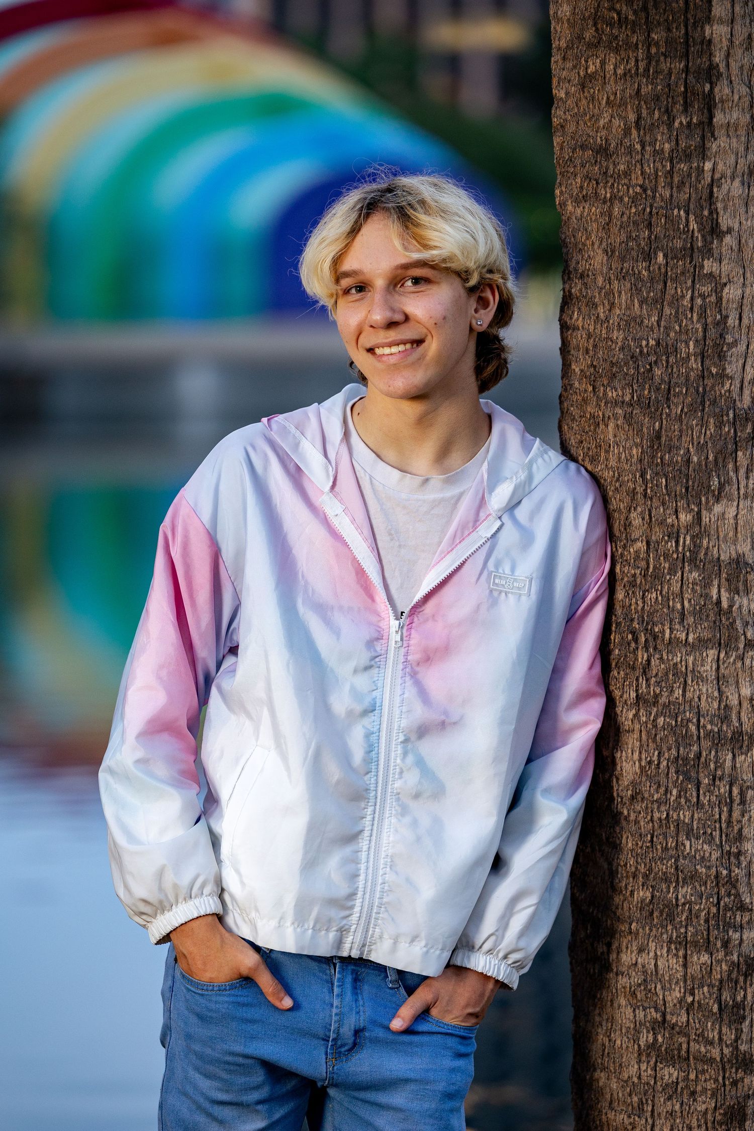 Smiling teen in pastel windbreaker leans against palm tree near colorful rainbow bridge outdoors.