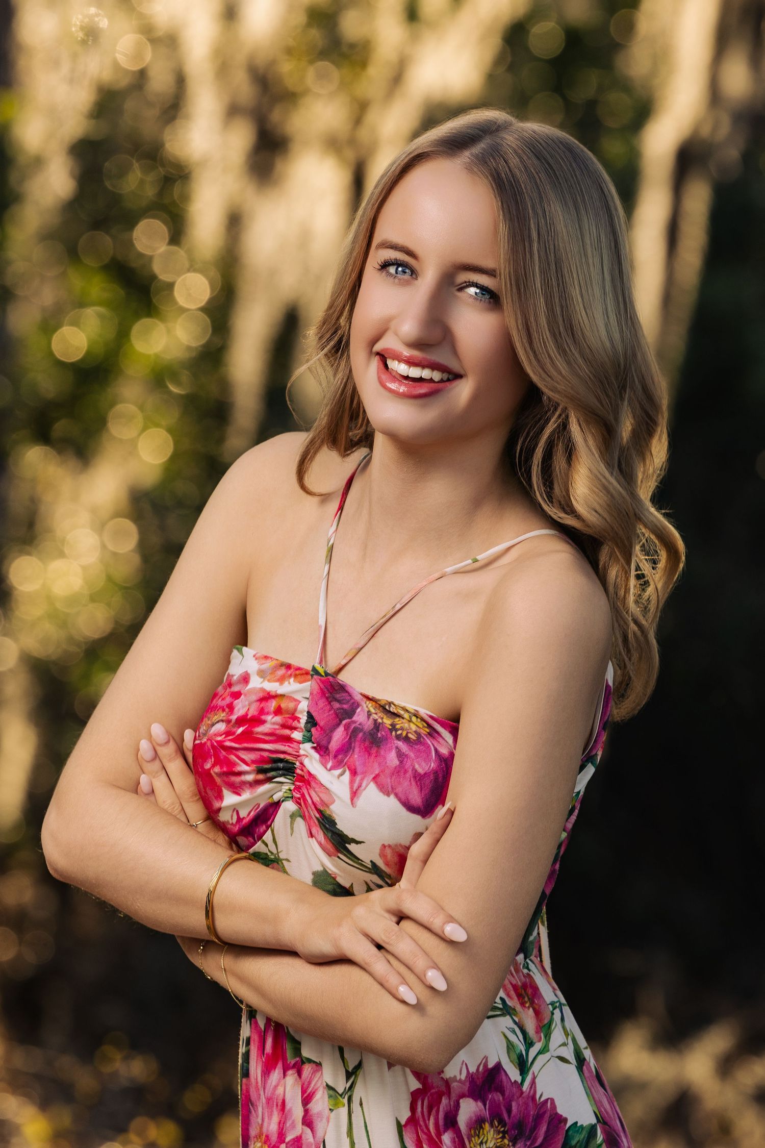 Smiling blonde woman in floral strapless dress with arms crossed outdoors.