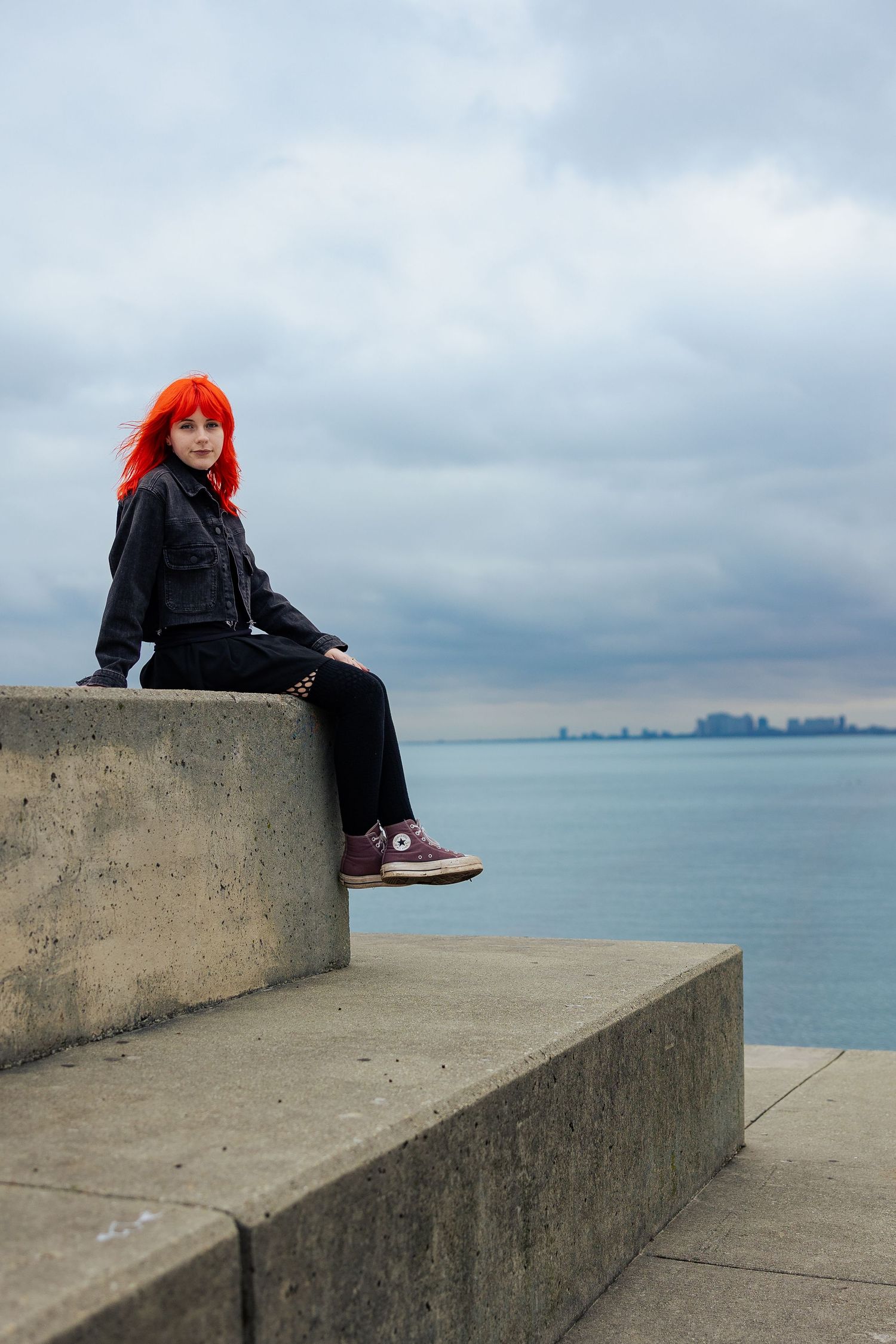 Red-haired woman in black outfit sitting on concrete steps by Lake Michigan on a cloudy day.
