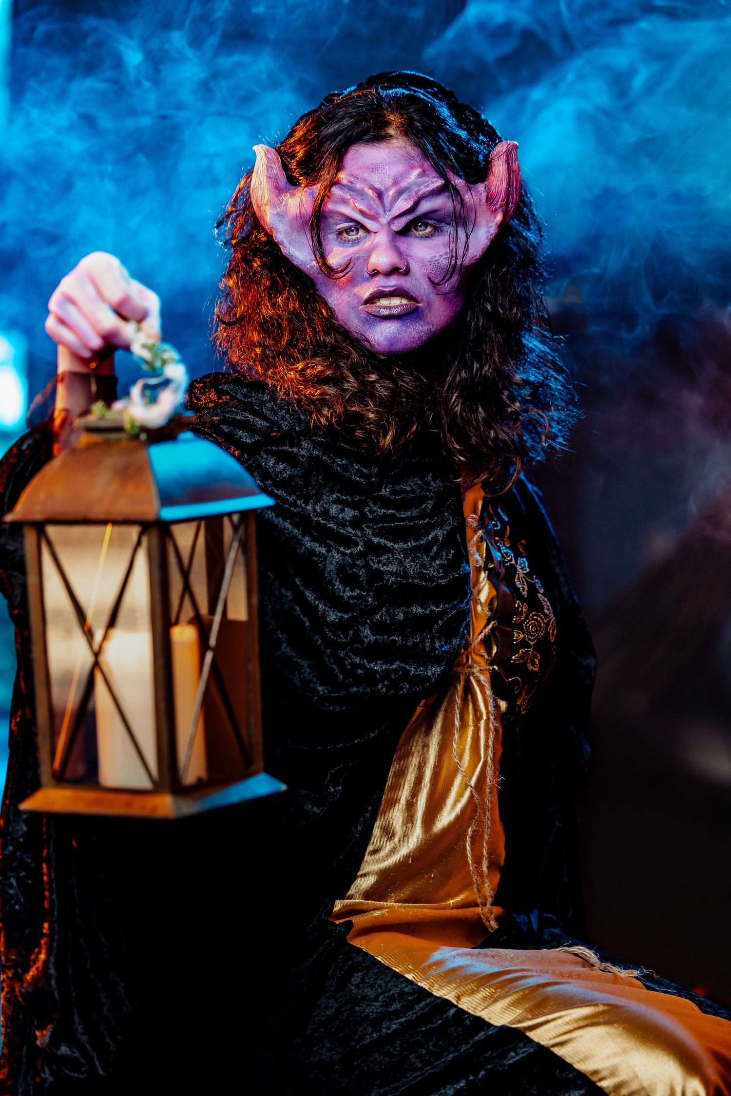 Menacing demon character with purple face paint and horns holds a glowing lantern in smoky blue light.