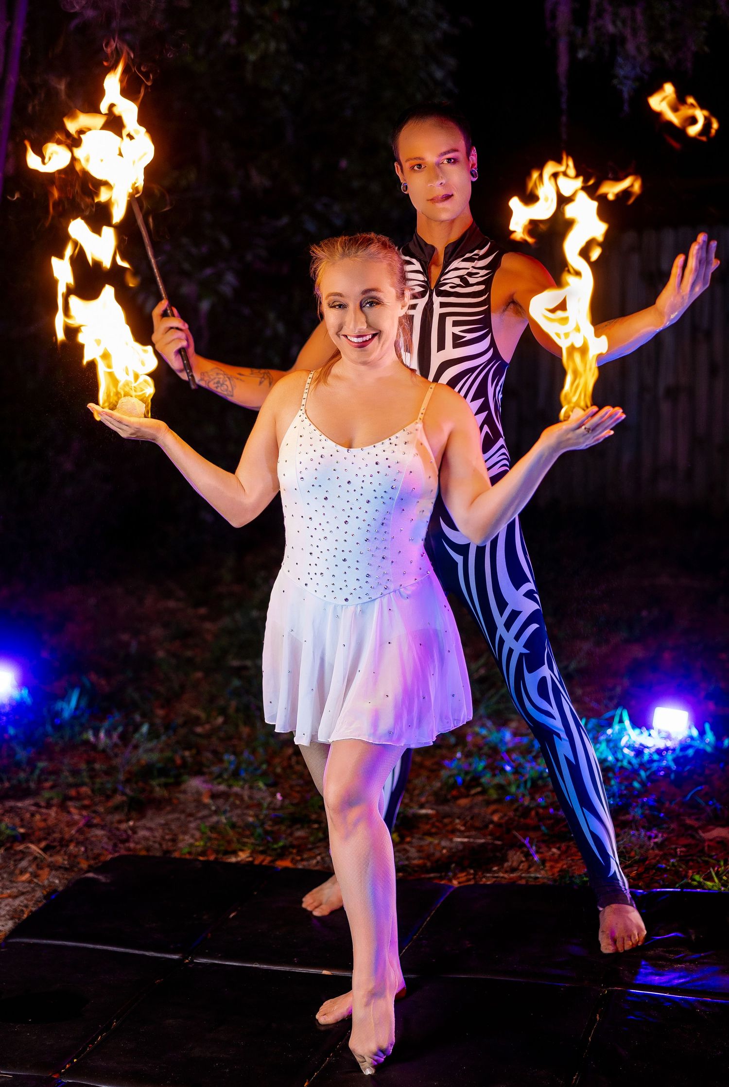 Dynamic duo of fire dancers perform outdoors at night, woman in white dress holding flaming torch with partner behind.