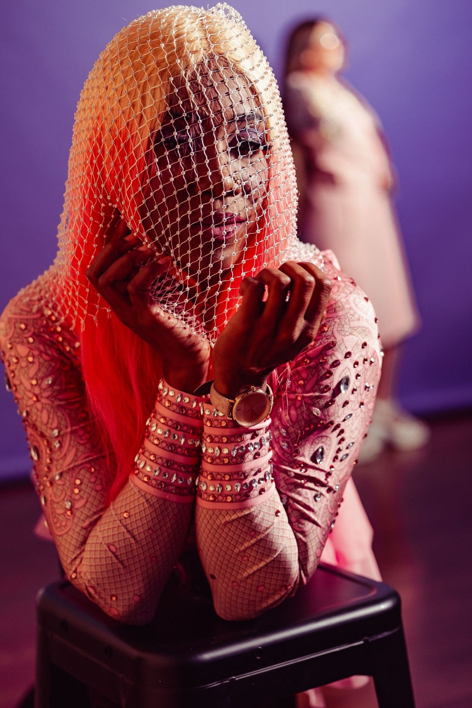 Woman in pink rhinestone bodysuit and mesh veil sits on stool under dramatic red lighting.