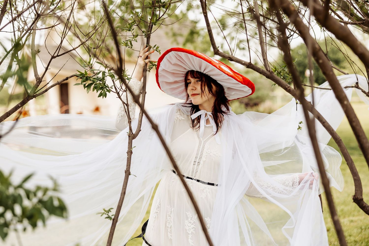 Woman in white dress and wide red-brimmed hat poses gracefully among tree branches outdoors.
