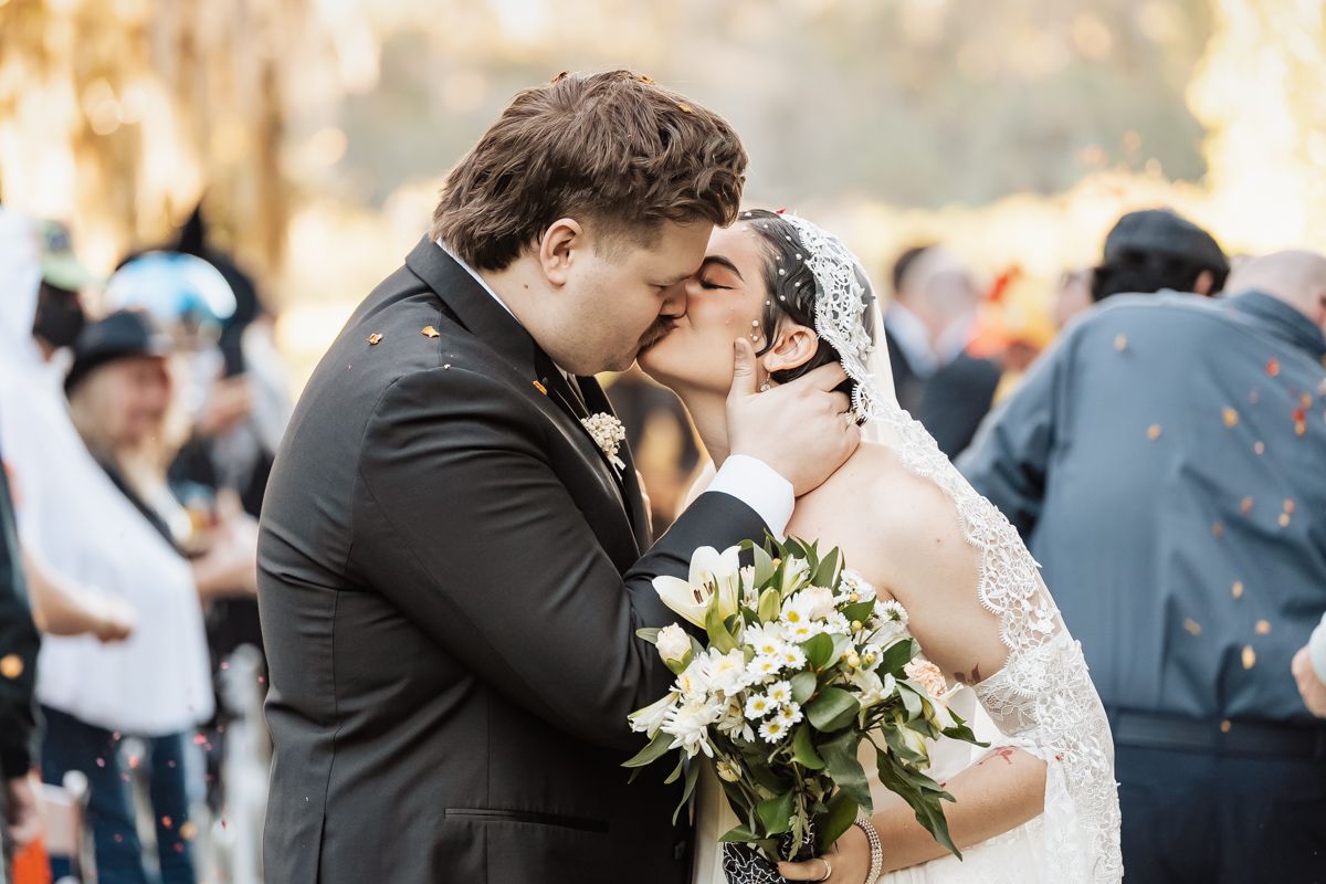 Bride and groom sharing first kiss while bride holds white floral bouquet at outdoor wedding.