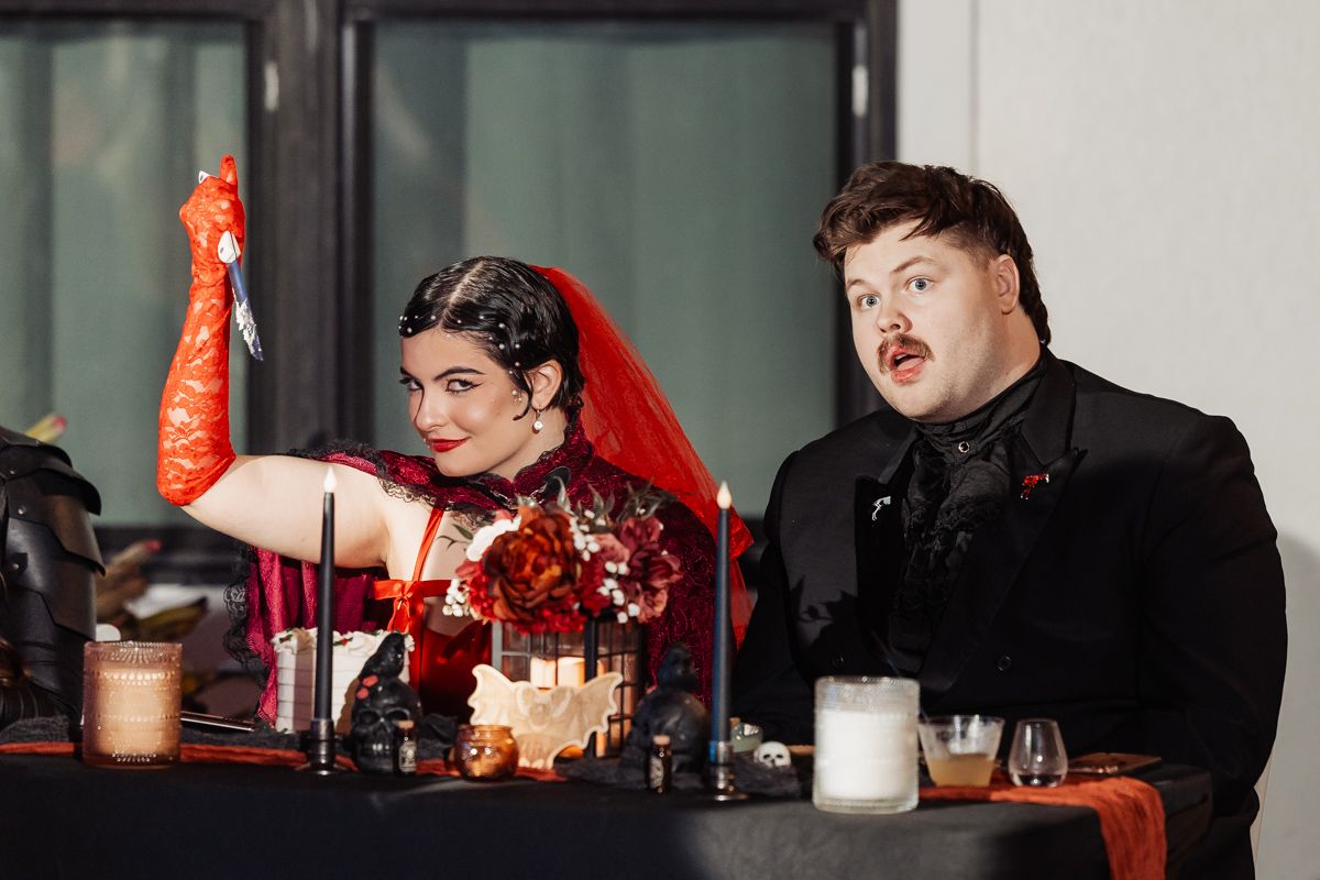 Gothic bride in red lace veil and groom in black suit seated at a Halloween wedding reception.