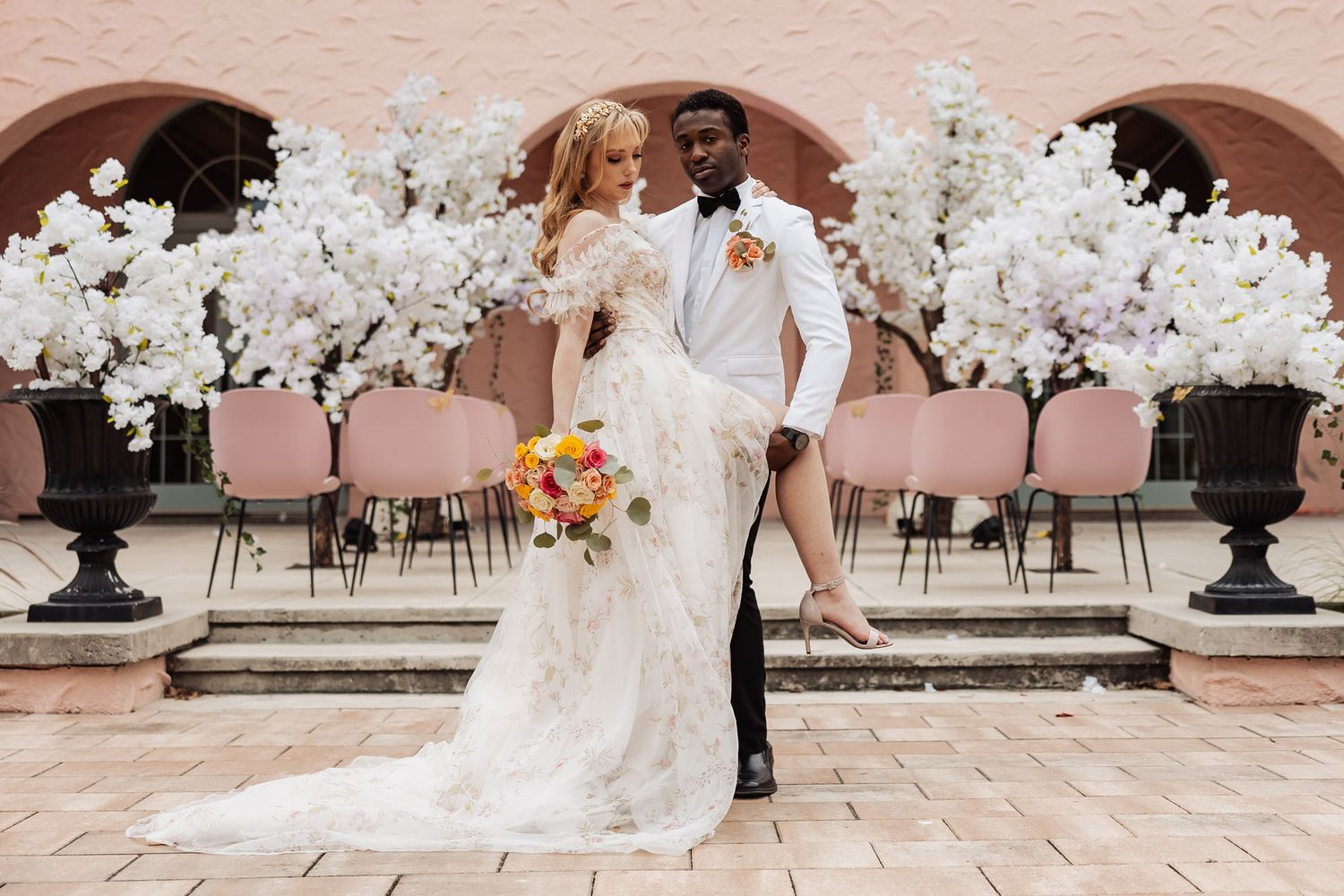 Interracial wedding couple posing elegantly before white cherry blossom trees in stunning venue.