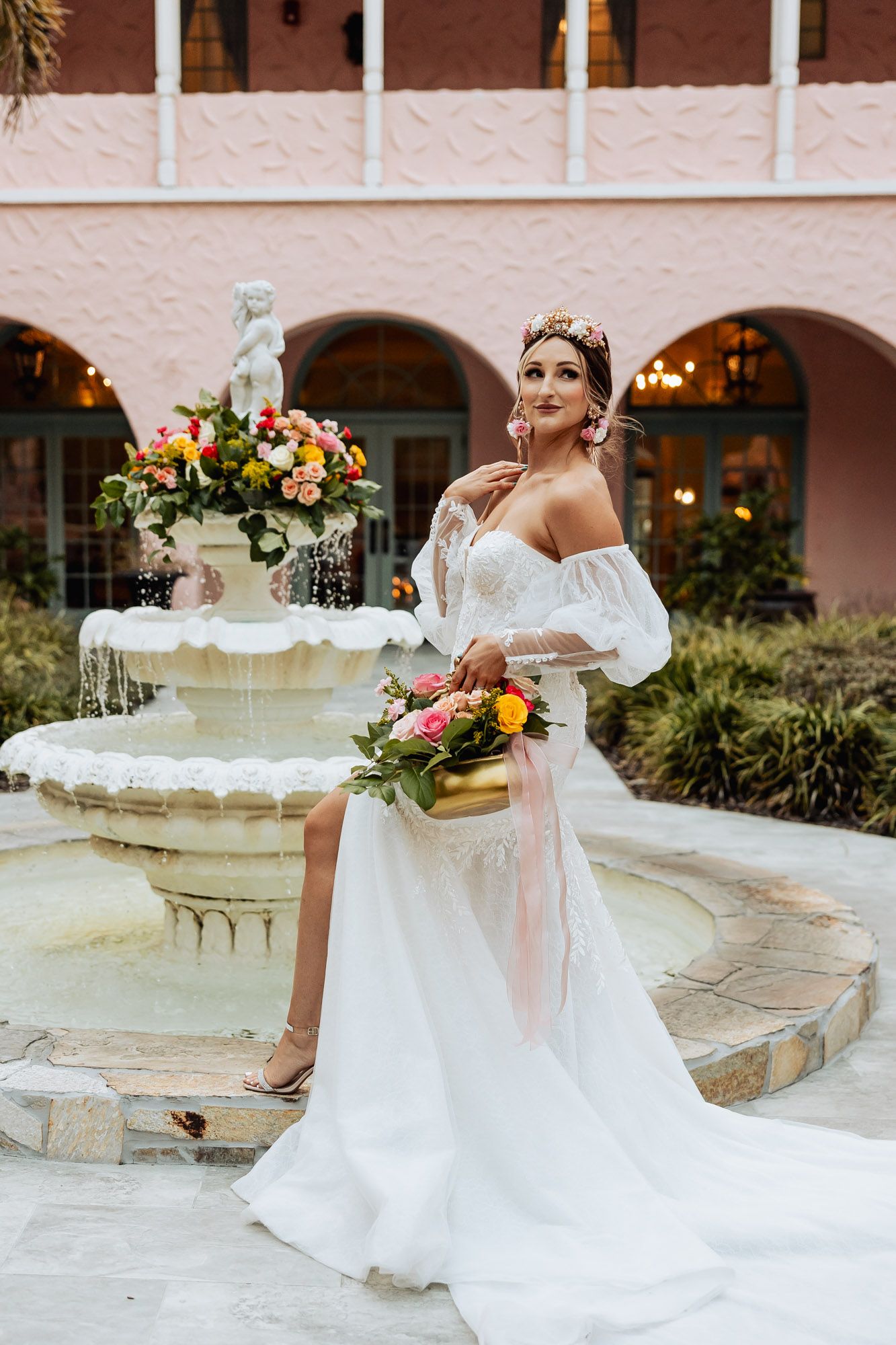 Bride in off-shoulder lace wedding dress poses by stone fountain holding vibrant floral bouquet at pink estate.