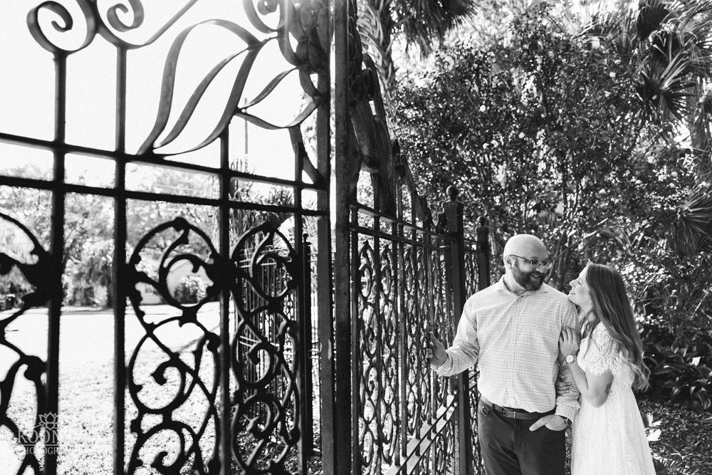 A couple poses together near an ornate wrought iron gate in a garden setting.