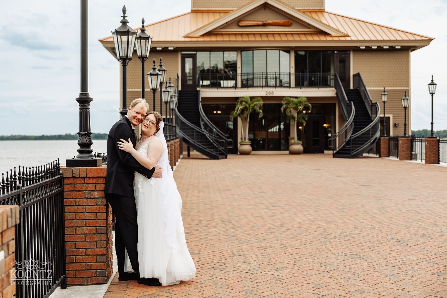 Newlyweds embrace outside waterfront venue with decorative iron railings and brick details.