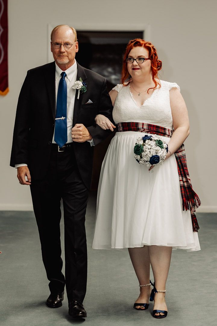 Bride with red hair in white dress with tartan sash walks down aisle with suited escort.