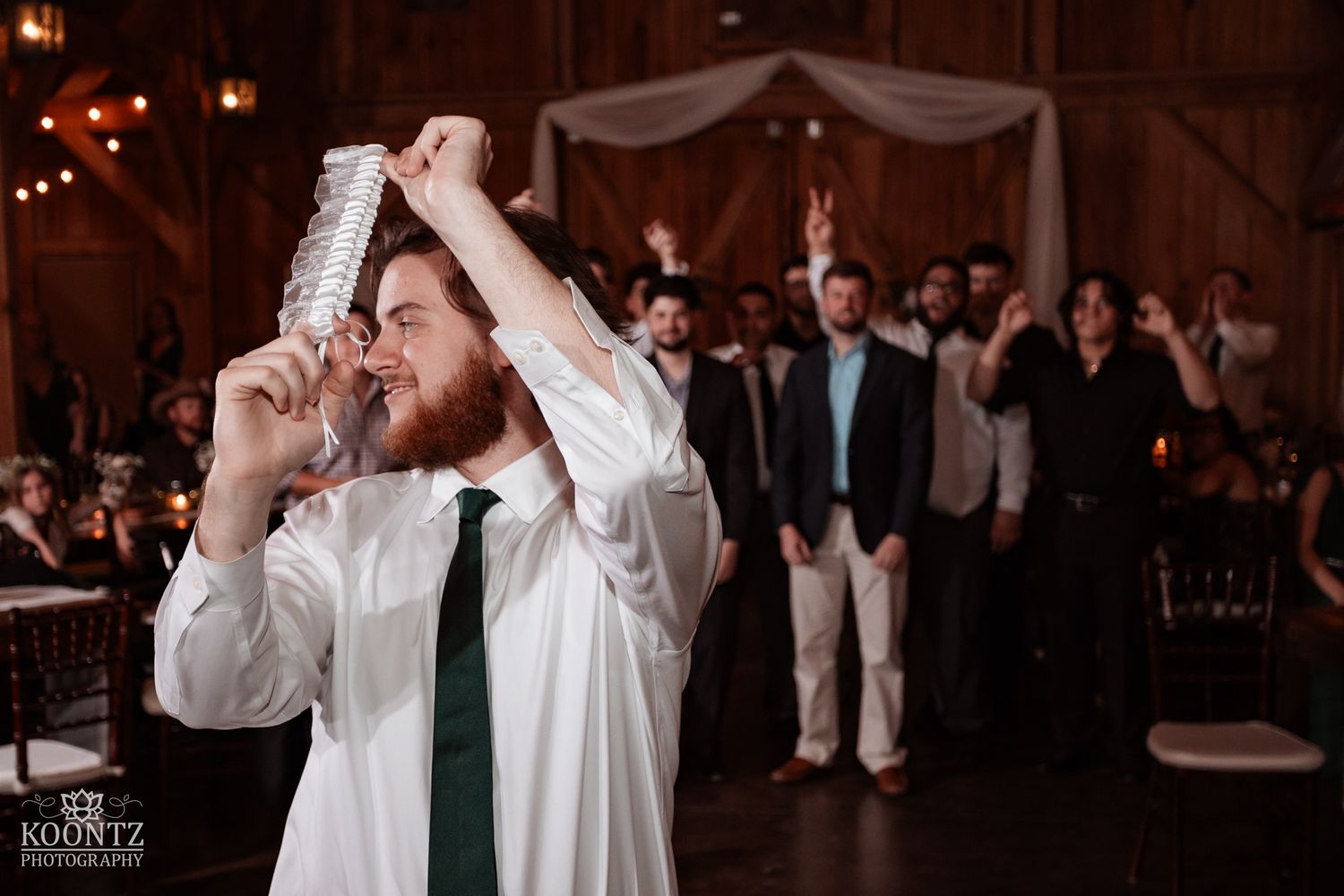 Excited groom drinks from a vase while celebrating at rustic barn wedding reception with guests.