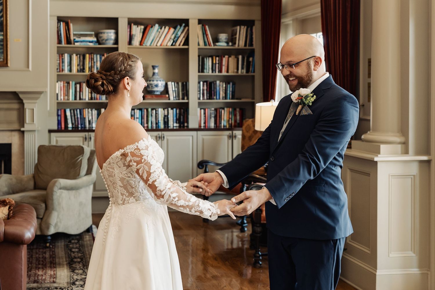 Bride and groom share intimate first look moment holding hands in elegant bookshelf-lined room.