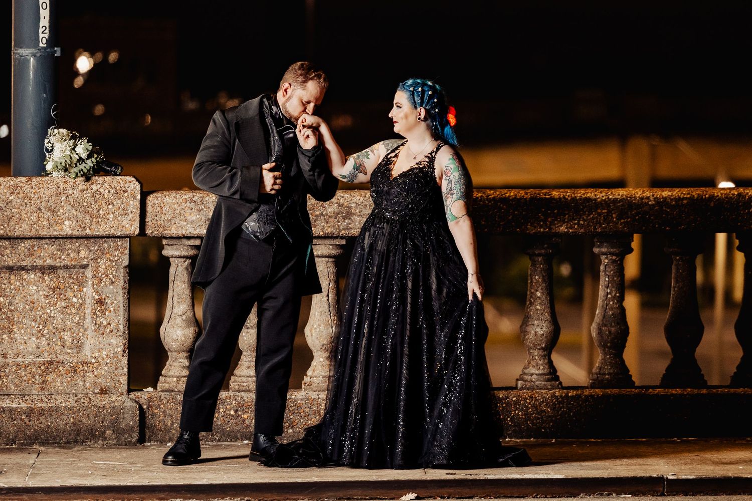 Gothic groom kissing bride's hand by stone balustrade at night outdoor wedding venue.