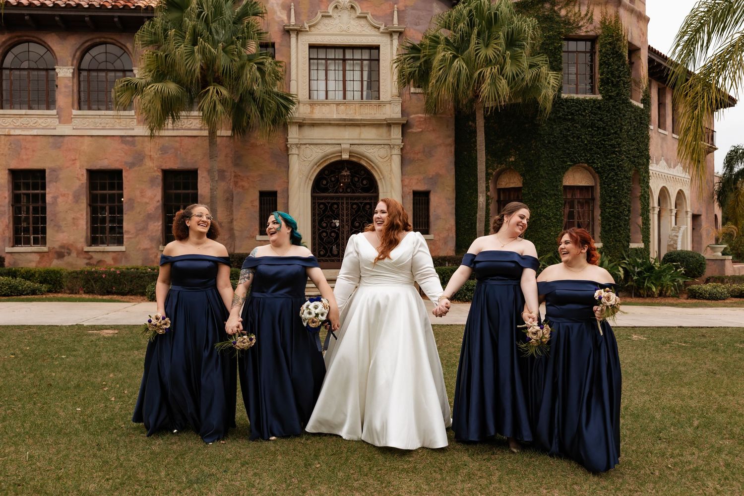 Bride in long-sleeve white gown holds hands with bridesmaids in navy dresses while strolling before a historic estate.