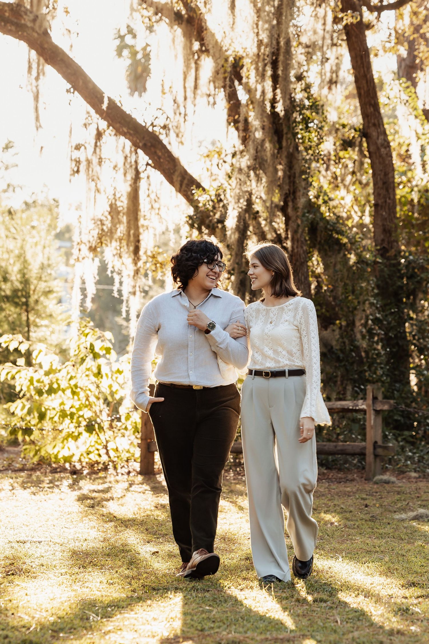 Two women walking together arm-in-arm beneath Spanish moss-covered trees in a sunlit park.