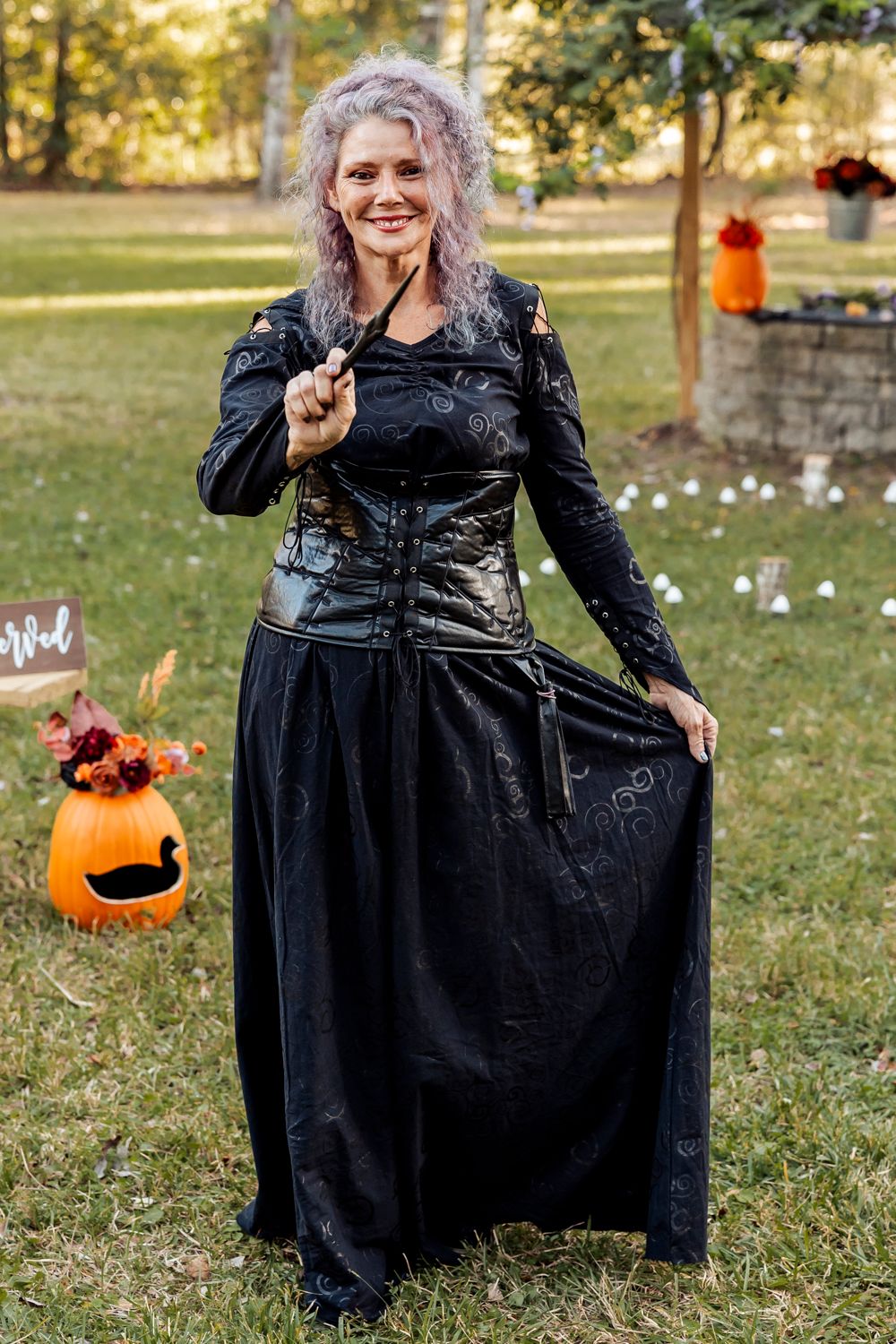 Person in black Victorian gothic dress poses with pumpkin decorations in an outdoor setting.
