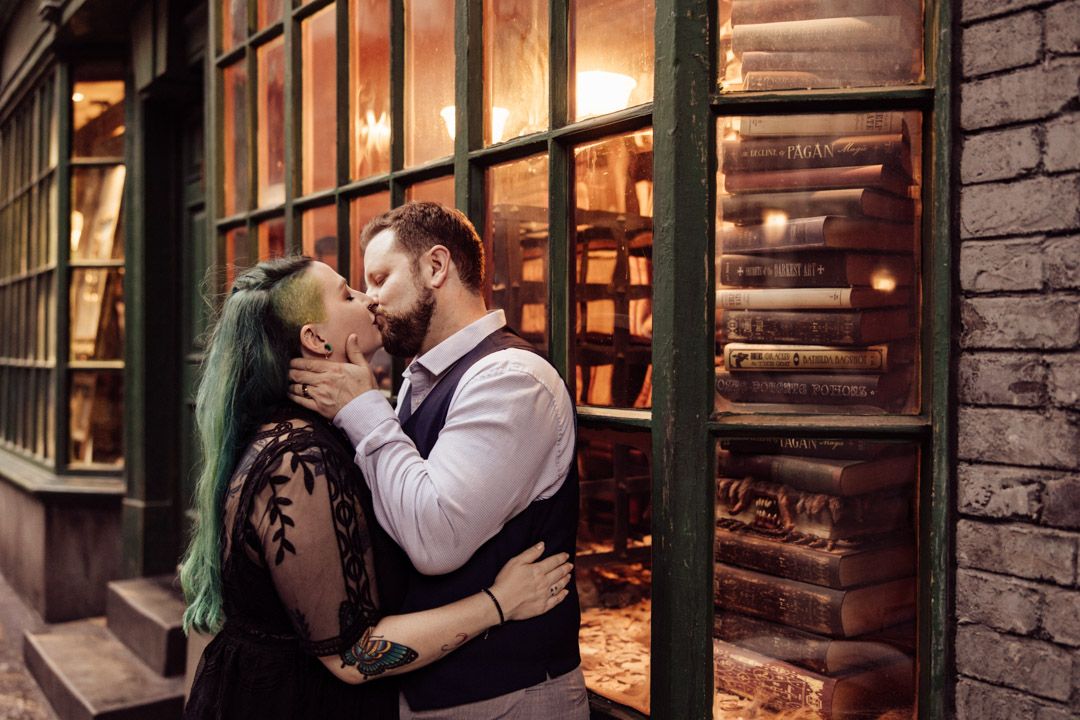 Couple sharing a romantic kiss in front of a warmly lit bookshop window filled with vintage books.