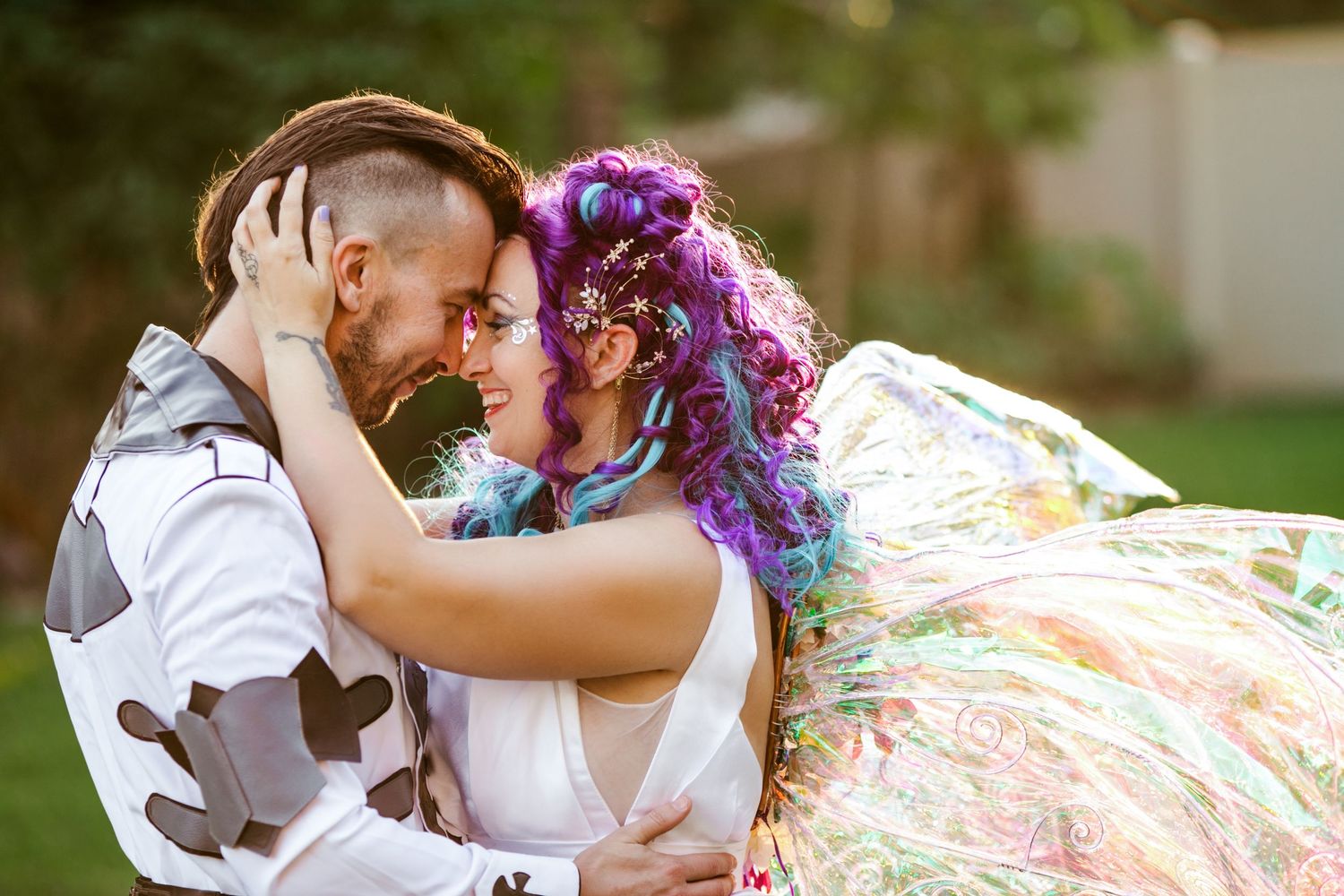 Couple in colorful wedding attire embrace outdoors, bride with purple hair and painted dress, groom in patterned suit.