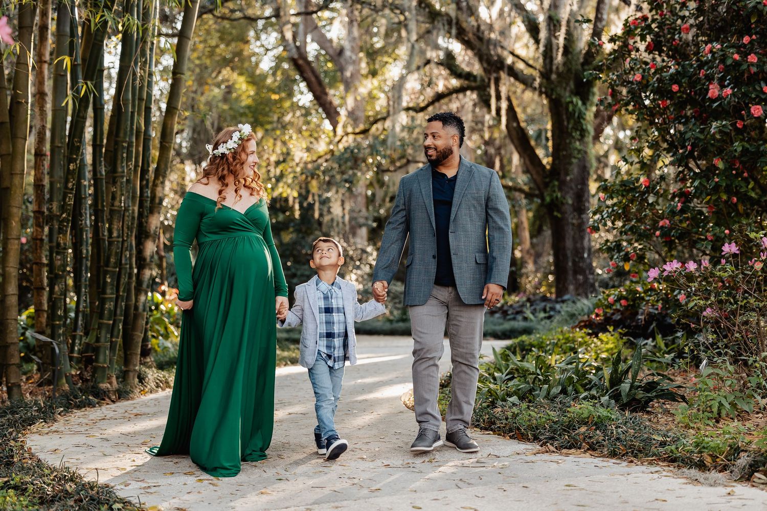 Pregnant woman in green gown with family on garden path surrounded by trees and pink flowers.