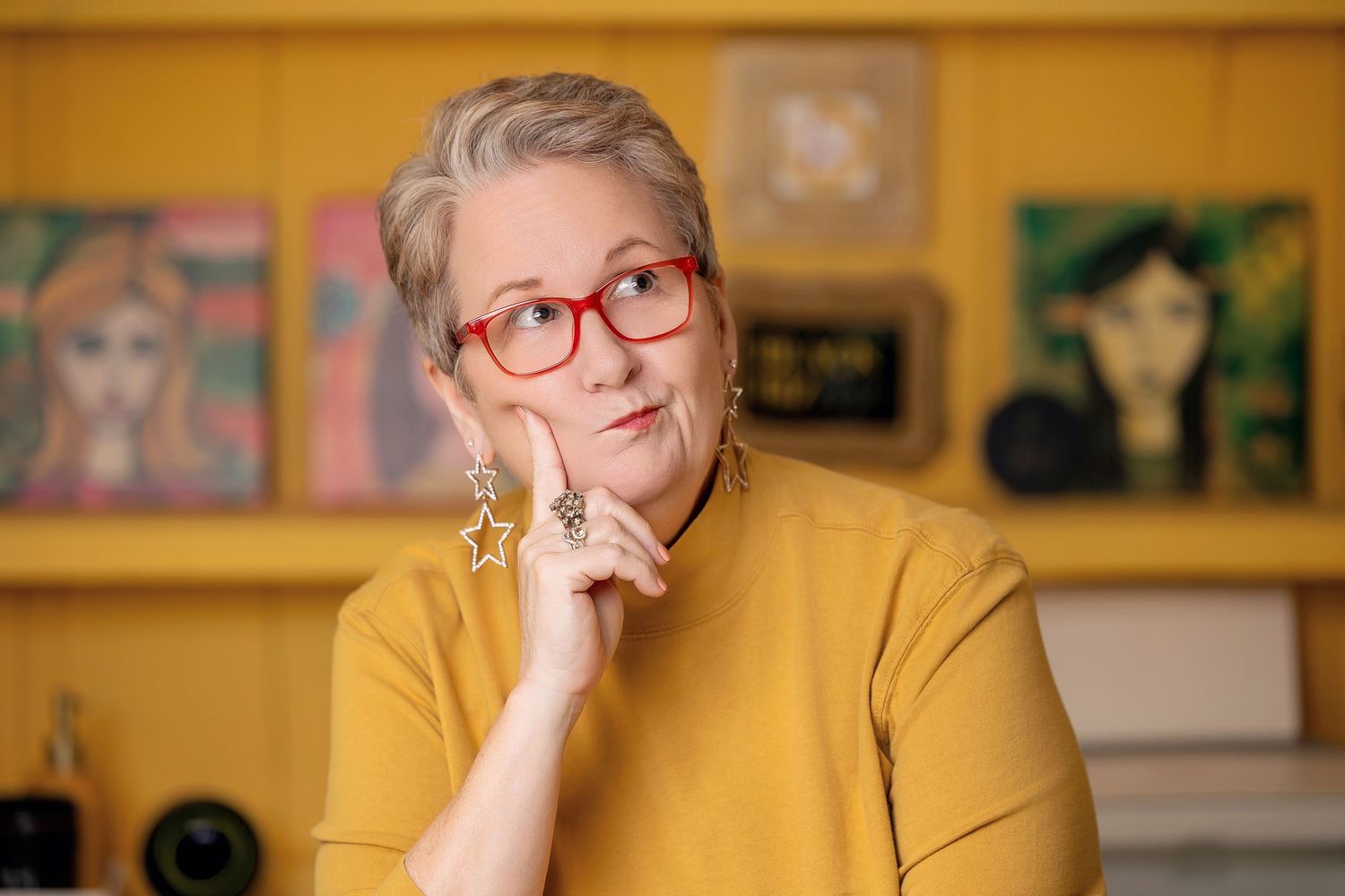 Thoughtful woman with red glasses and yellow top rests chin on hand in a creative office environment.