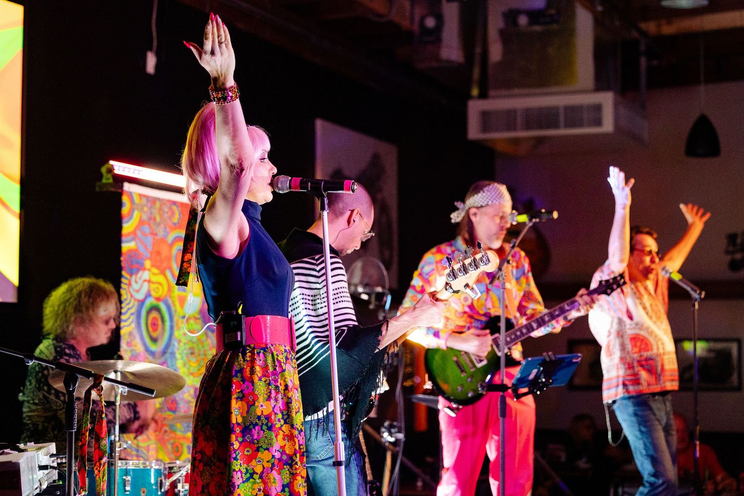 Energetic band performing on stage in vibrant floral outfits with psychedelic backdrop projection.