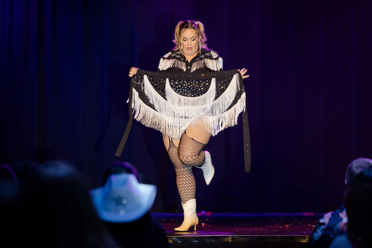Burlesque dancer in black fringe western costume lifting leg on stage platform.