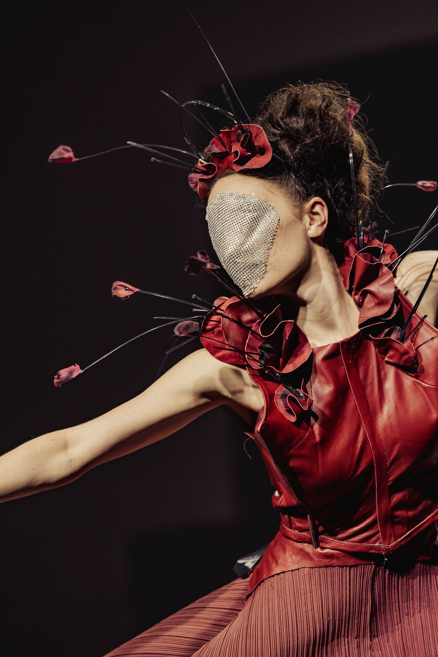 Close-up of dancer in red leather corset with floral headpiece and silver mesh face mask mid-performance.