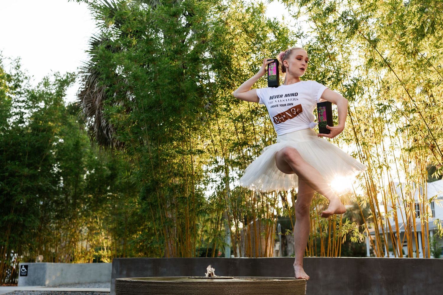 Ballerina in white tutu holding bottles while dancing en pointe near bamboo trees outdoors.