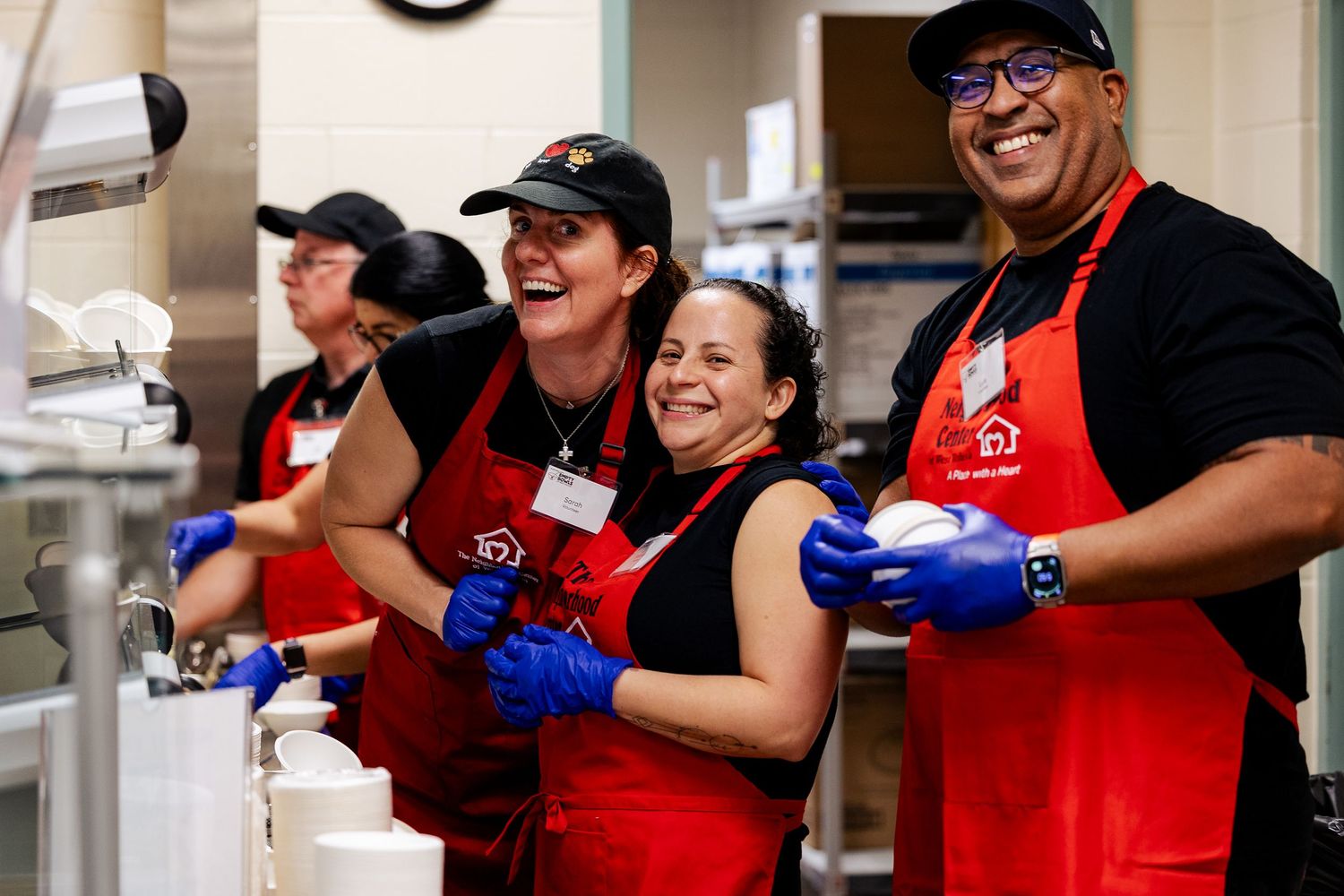 Three joyful volunteers in red aprons pose together while serving food in a community kitchen.
