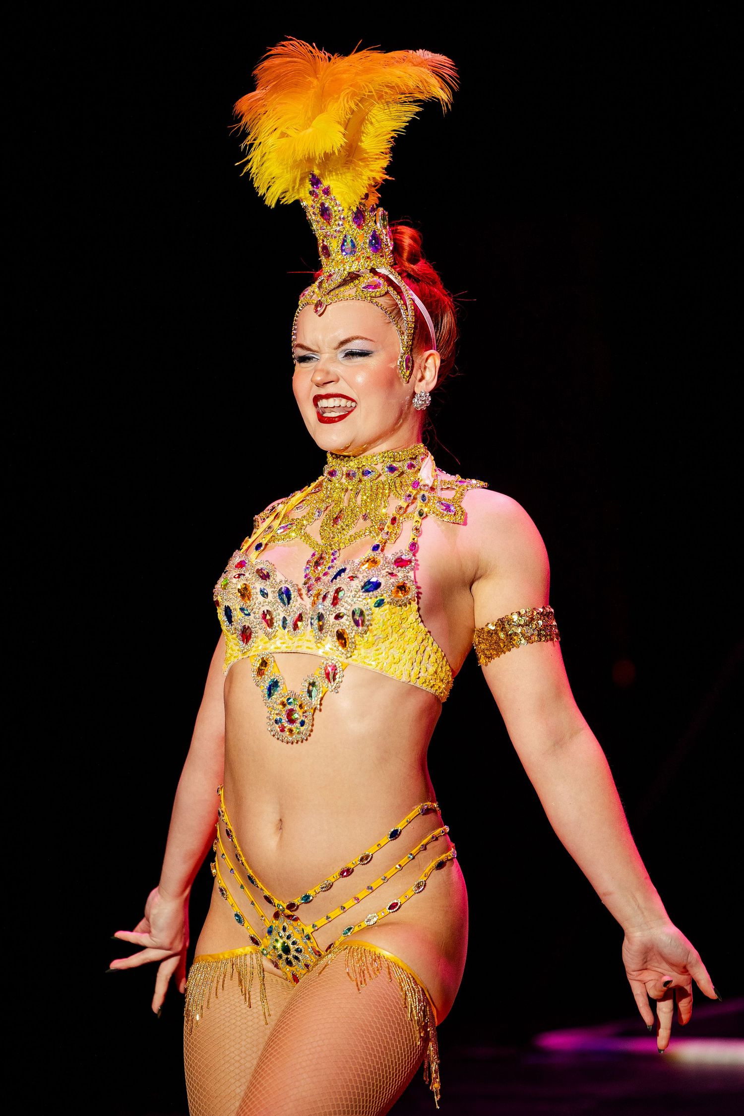 Burlesque performer in jeweled golden bikini costume with orange feather headdress smiles on dark stage.