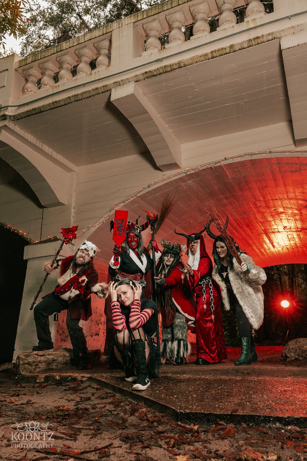 Group of cosplayers in dark fantasy devil costumes pose under a bridge with dramatic red lighting.