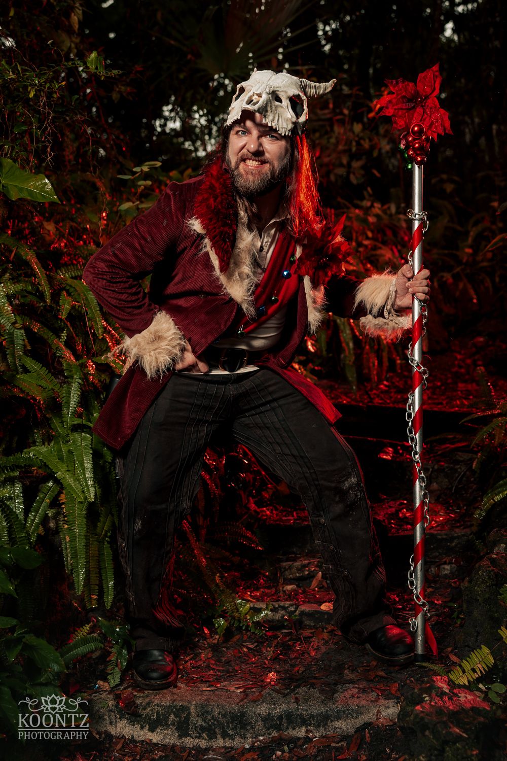 A bearded man in dark fantasy cosplay with animal skull hat, red fur coat, and ornate red staff poses in a forest.