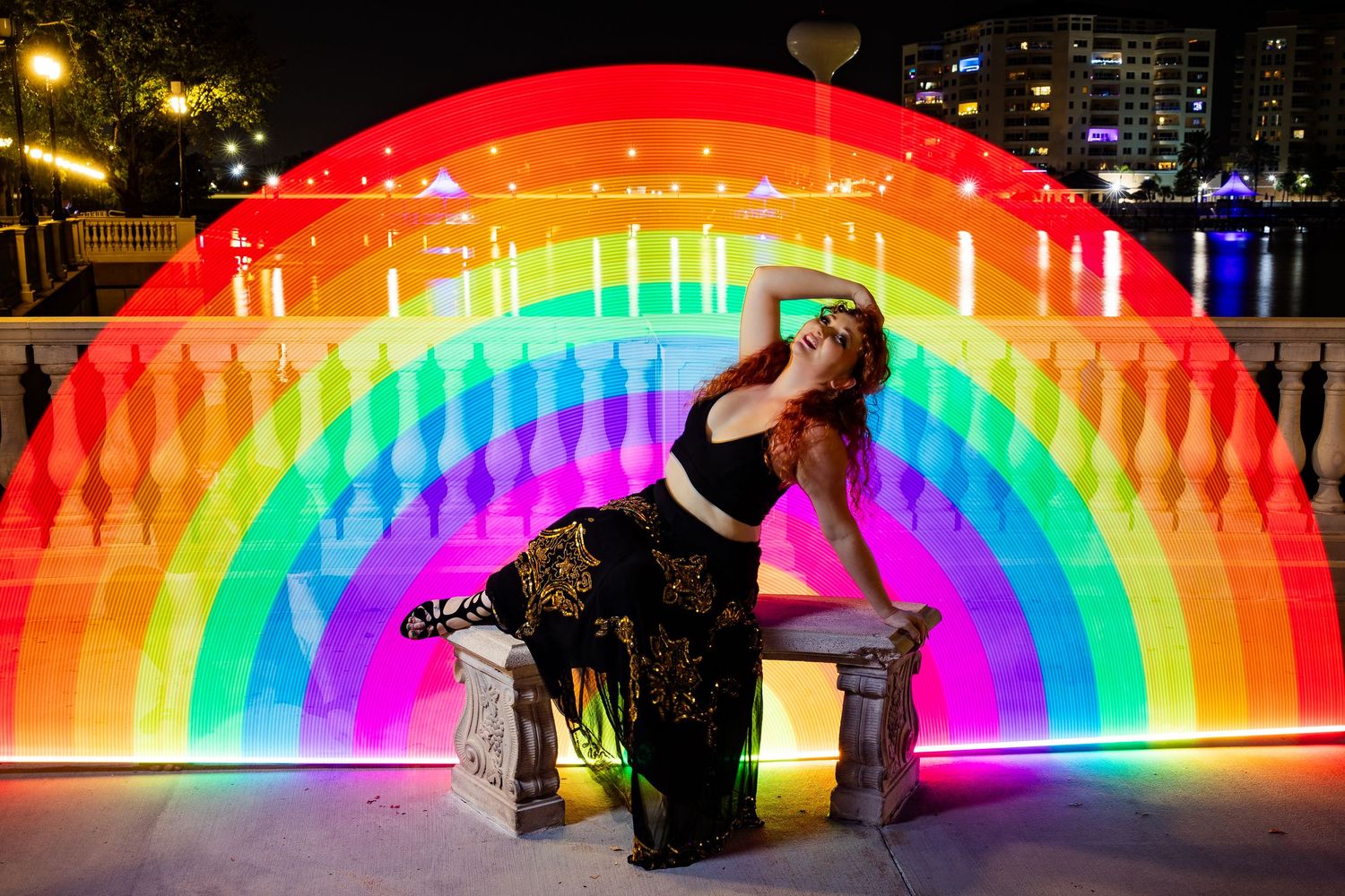 Woman in black dress poses dramatically under a vivid rainbow arch light installation at night.