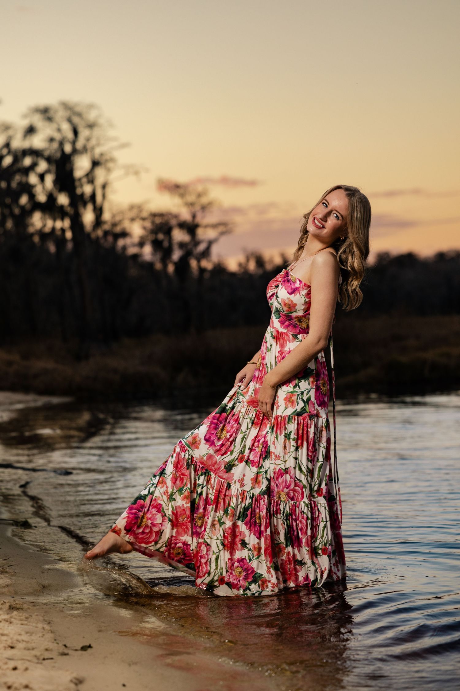 Smiling blonde woman in flowing floral maxi dress wading in water at dusk.