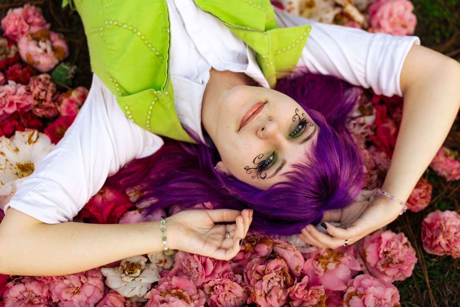 Woman with purple hair and green eye makeup lying among pink roses and flowers outdoors.