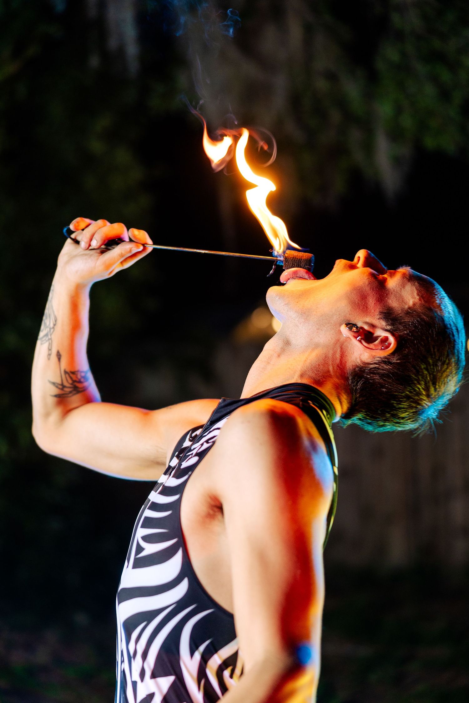Fire performer in tribal costume breathes fire from a torch during an outdoor nighttime circus show.