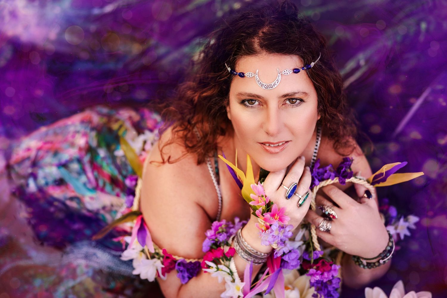 Bohemian woman with jeweled headpiece and floral accessories poses against a vibrant purple backdrop.