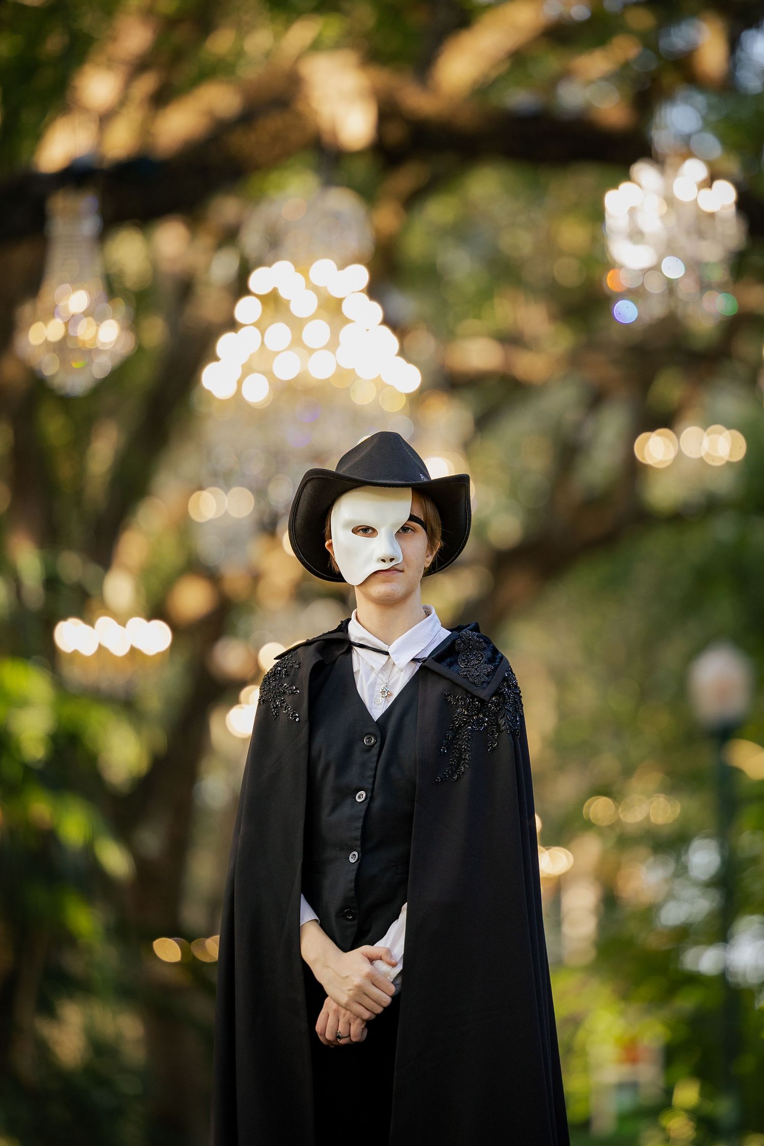 Person in Phantom of the Opera costume with white mask, black cape, and cowboy hat standing outdoors with bokeh lights.