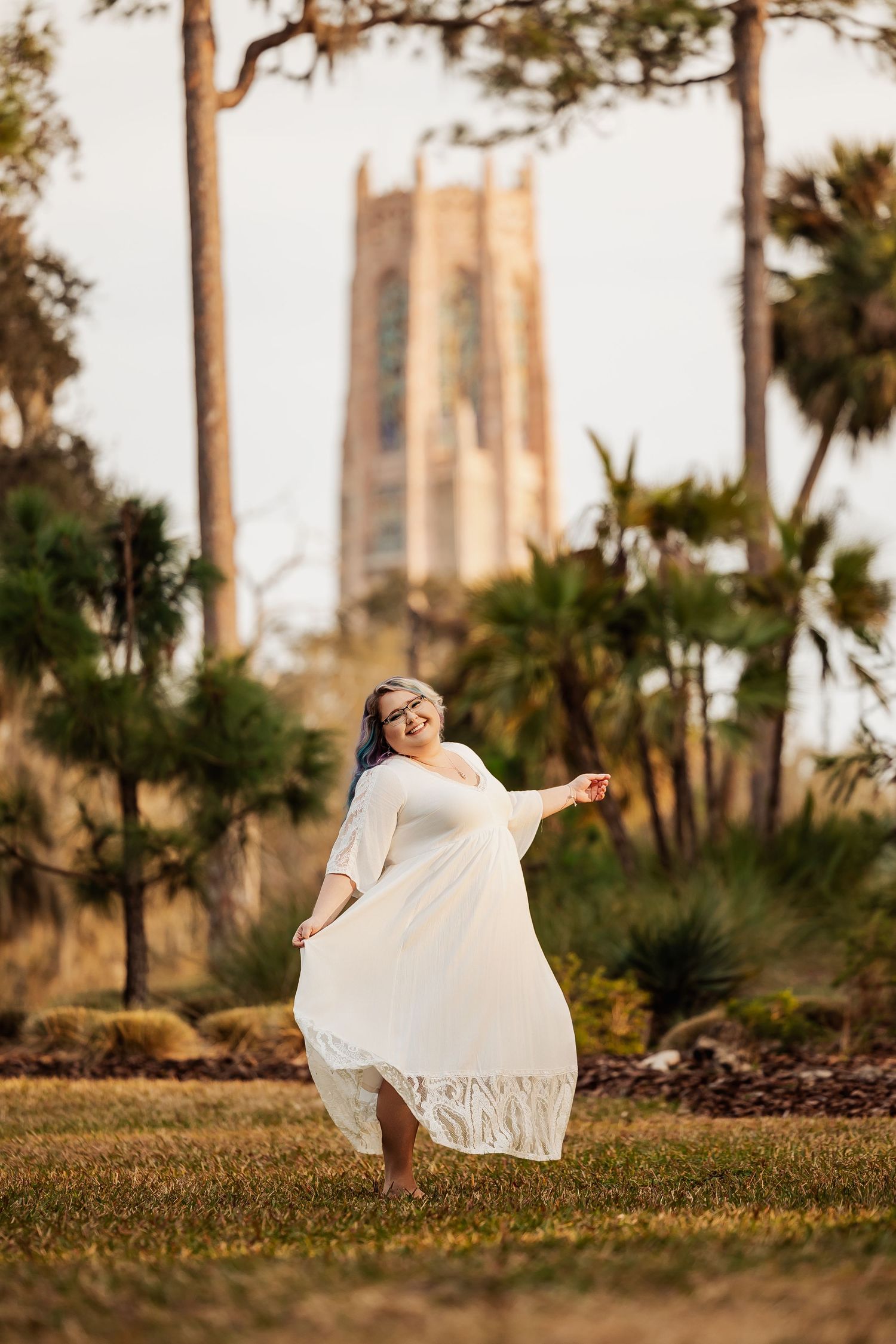 Woman twirls in white lace dress on grass with tall tower and palm trees behind her.