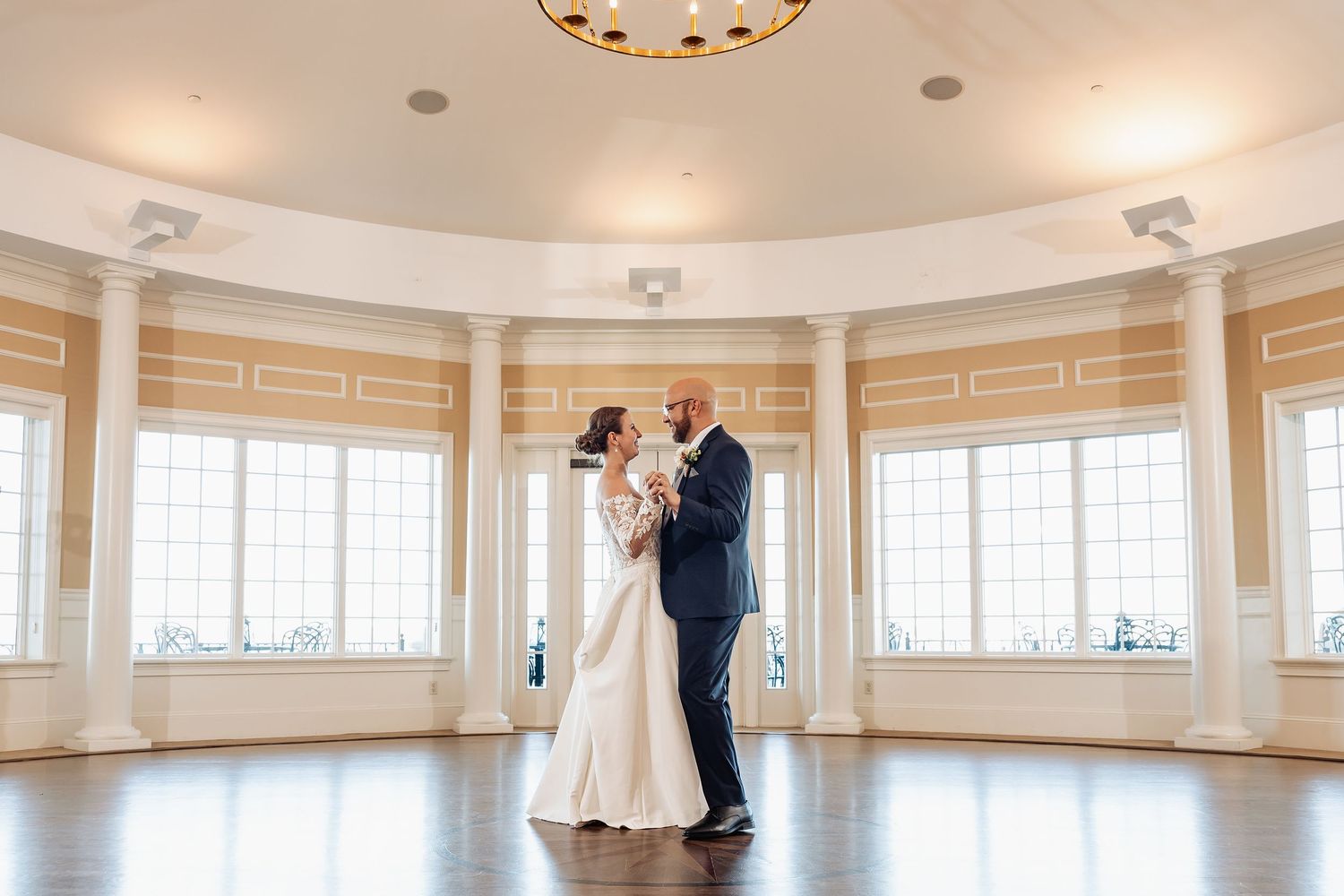Bride and groom embrace during first dance in stunning circular ballroom with floor-to-ceiling windows.