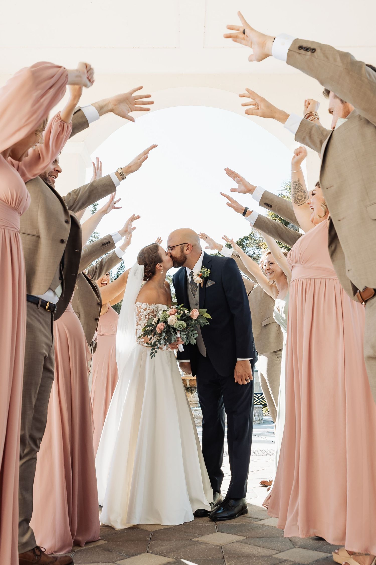 Newlyweds share a kiss under an archway as wedding party members raise their hands in celebration.