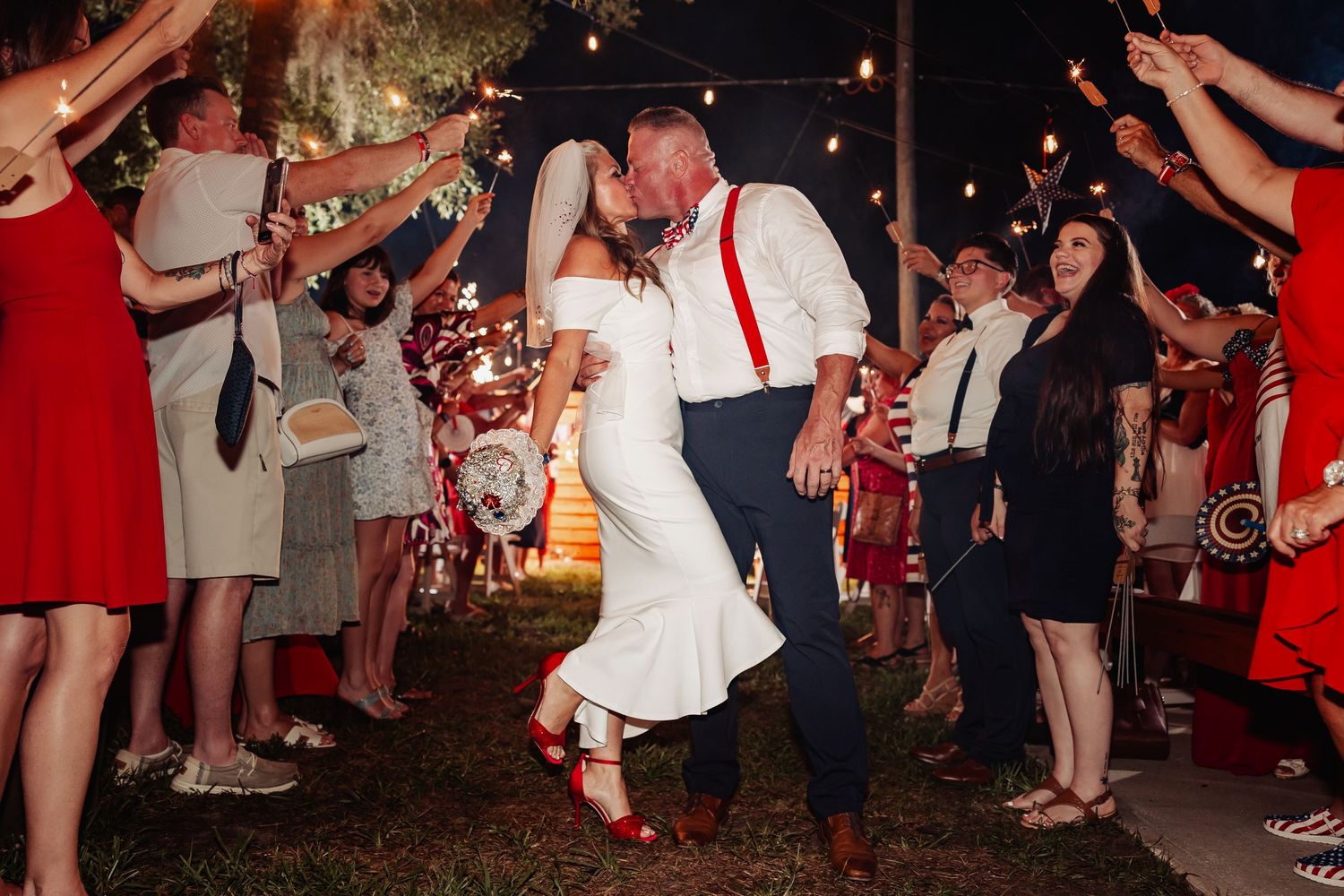 Bride and groom share romantic kiss during sparkler exit with cheering guests lining the outdoor wedding path.