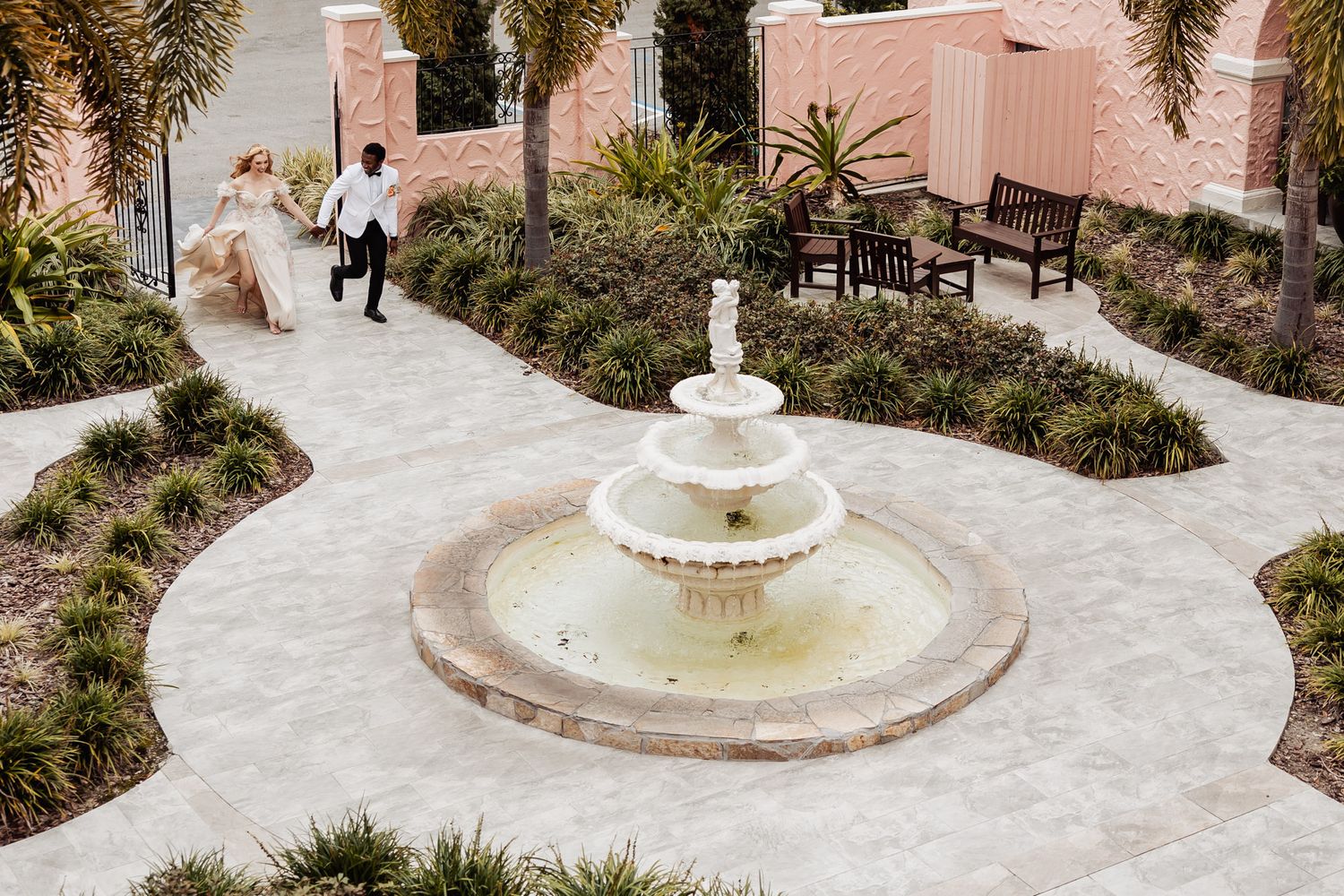 Aerial view of bride and groom walking past elegant white fountain at pink venue courtyard.