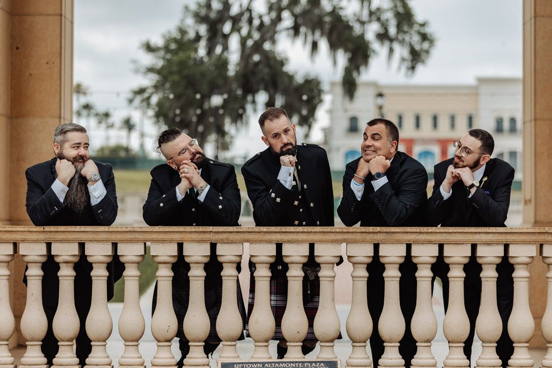 Groomsmen in formal attire leaning on a balcony railing making playful expressions.