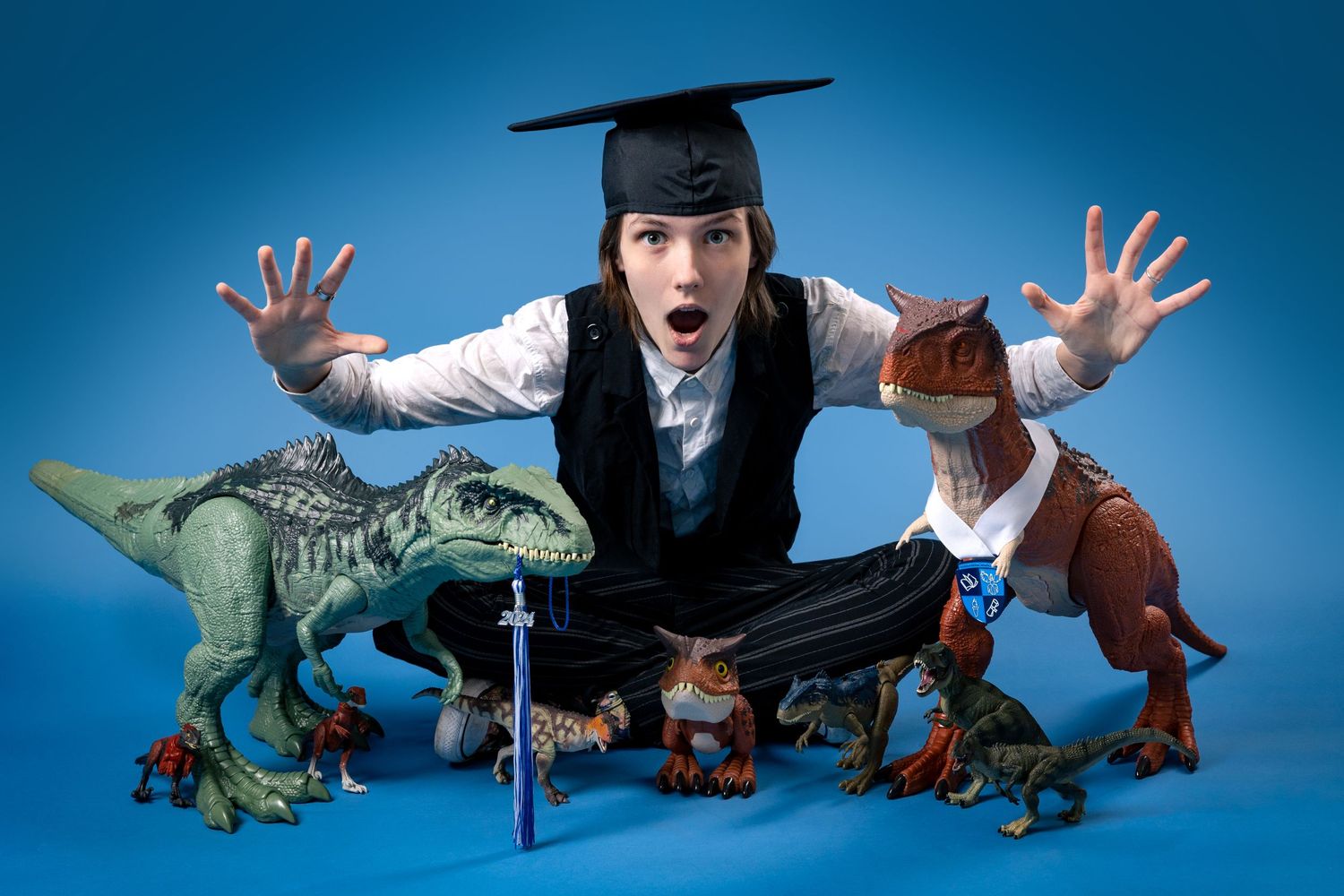 Playful graduation photo with person in cap and gown posing with toy dinosaurs against blue backdrop.
