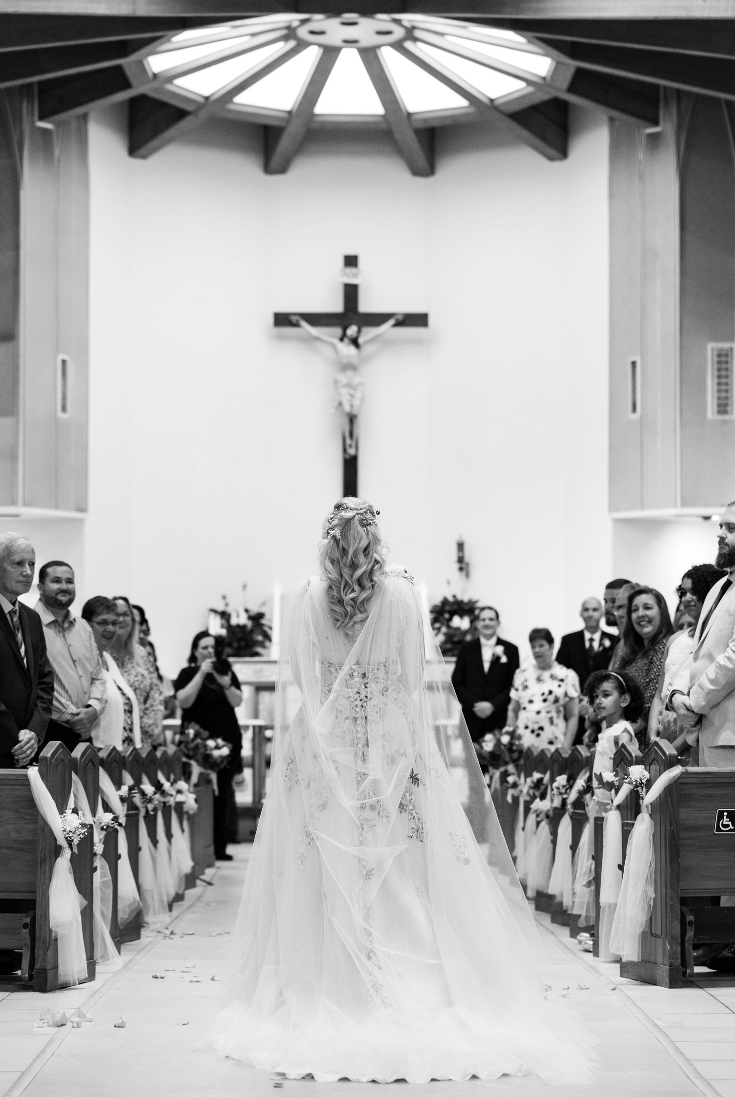 Black and white photo of bride walking down church aisle toward groom beneath crucifix.
